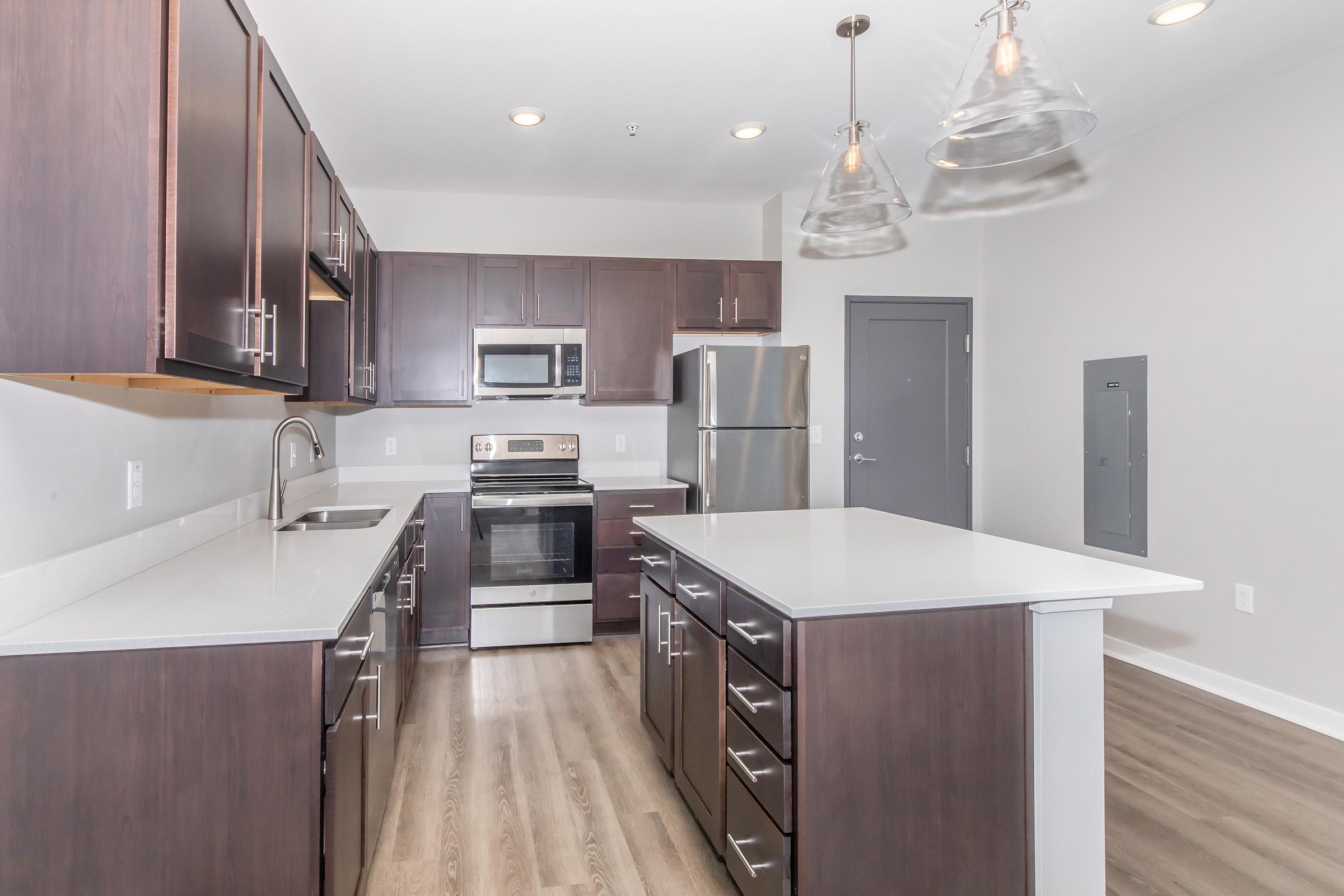 Modern kitchen featuring dark wood cabinetry, stainless steel appliances, a large island with a white countertop, pendant lighting, and hardwood floors. The kitchen includes a built-in microwave, a refrigerator, an oven, and a neutral-colored wall, creating a contemporary and open design.