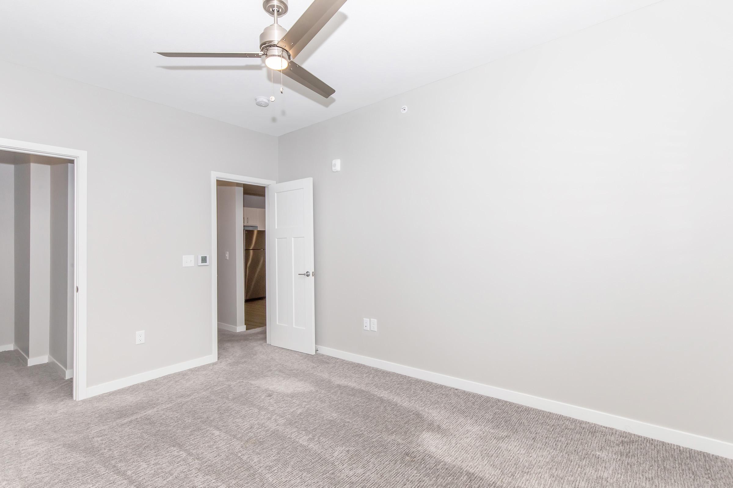 A spacious, empty room with light gray walls and carpet flooring. A ceiling fan is installed, and there's a doorway leading to another room. The door is white with a modern design, and the lighting creates a bright and inviting atmosphere.