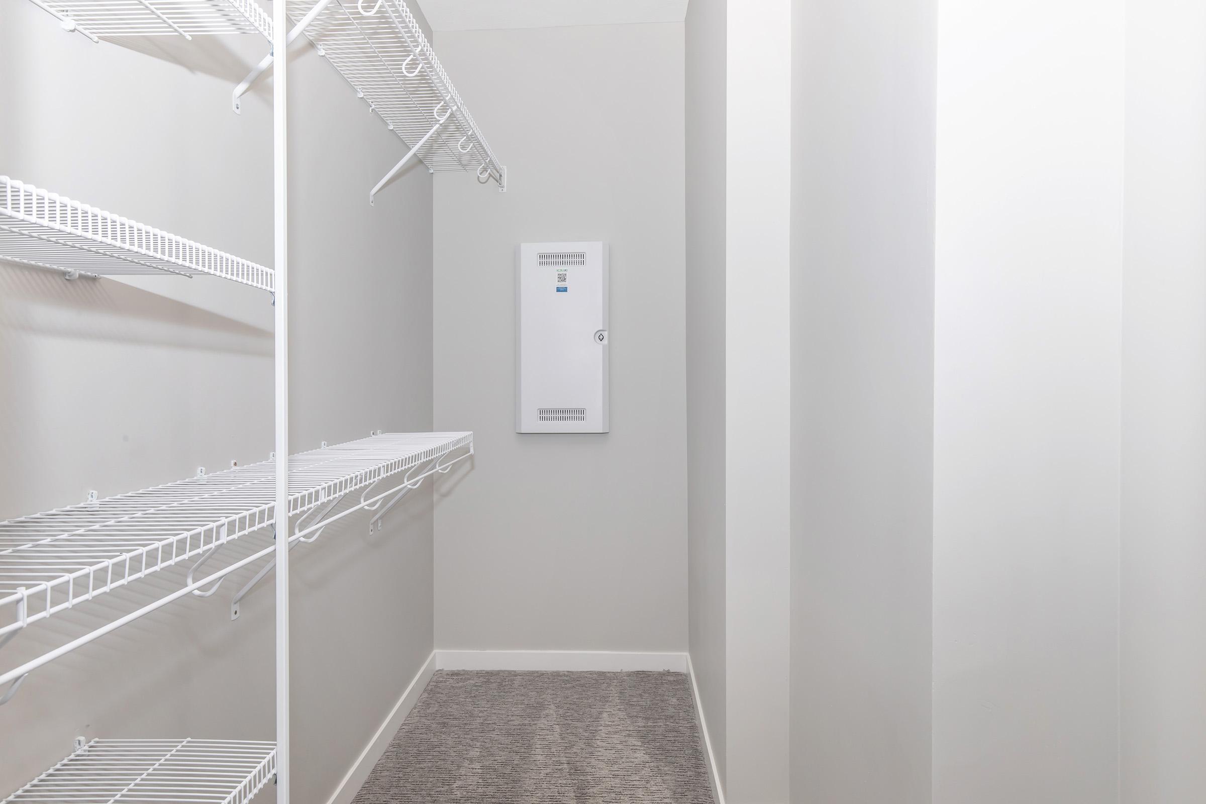 A spacious walk-in closet with light gray walls, featuring metal wire shelving along one side. The opposite wall has a white electrical panel, and the floor is covered with a plush gray carpet. Natural light brightens the area, creating an organized and airy feel.