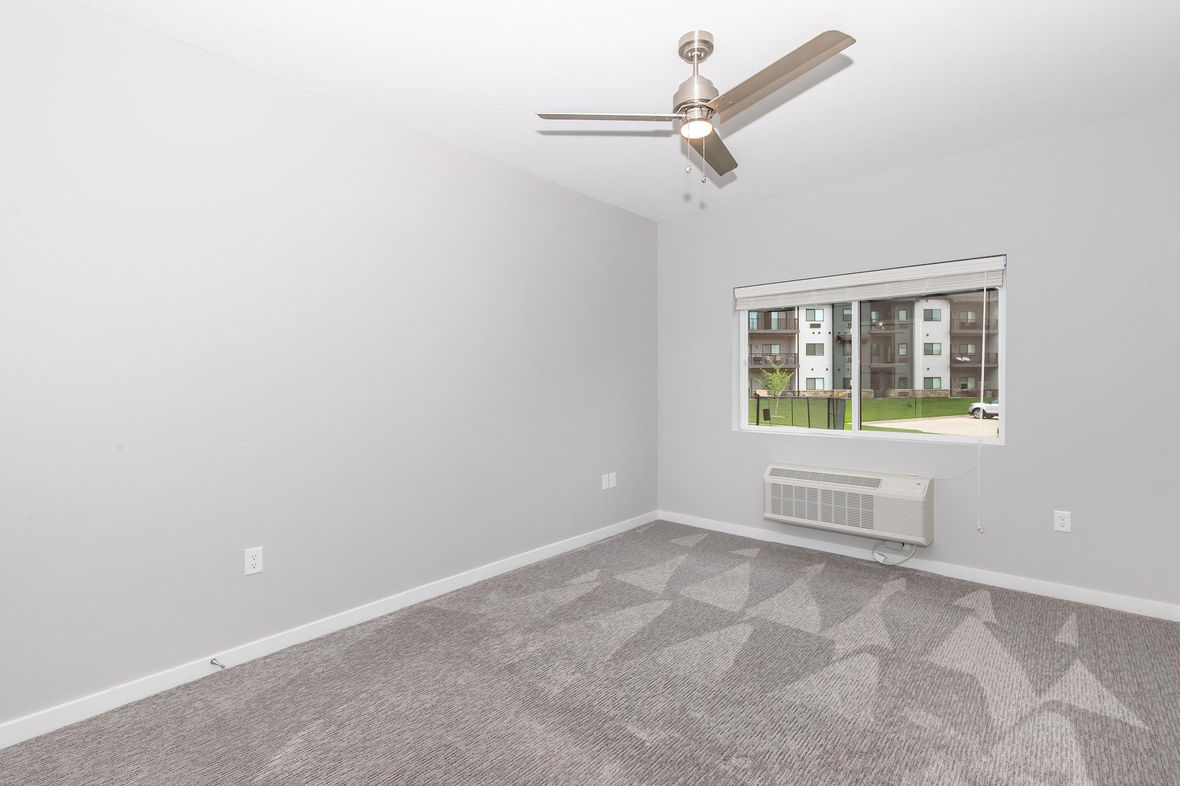 A spacious, empty room with light gray walls and a carpeted floor featuring a geometric pattern. A ceiling fan is installed, and there is a large window with a view of green outdoor space. An air conditioning unit is mounted below the window, providing a modern touch to the minimalist decor.