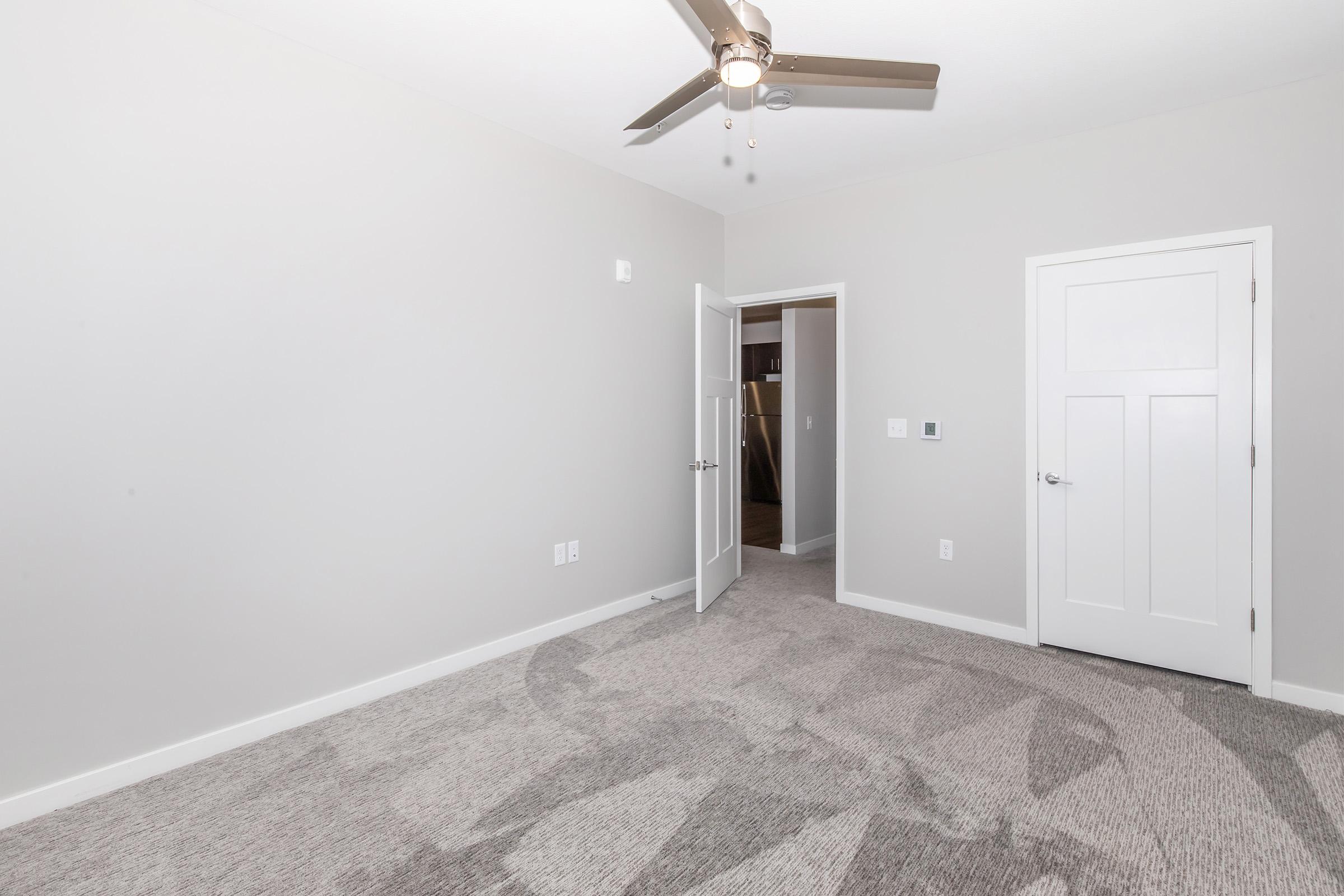 A spacious, neutral-colored room with light gray walls and a ceiling fan. The floor is covered with soft carpeting featuring a subtle pattern. There is an open door leading to another space, and a closed door on the right side, indicating a closet or bathroom. The room is well-lit and empty, providing a blank canvas.