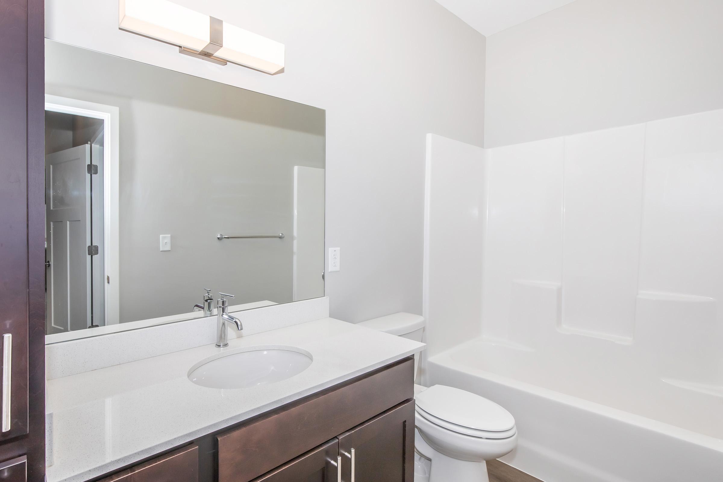 A modern bathroom featuring a white sink with a chrome faucet, dark wood cabinetry, a large mirror, and a neutral-colored wall. There is a white bathtub and shower combo, a toilet, and a towel rack on the wall. The space is well-lit with a fixture above the mirror, creating a clean and contemporary look.