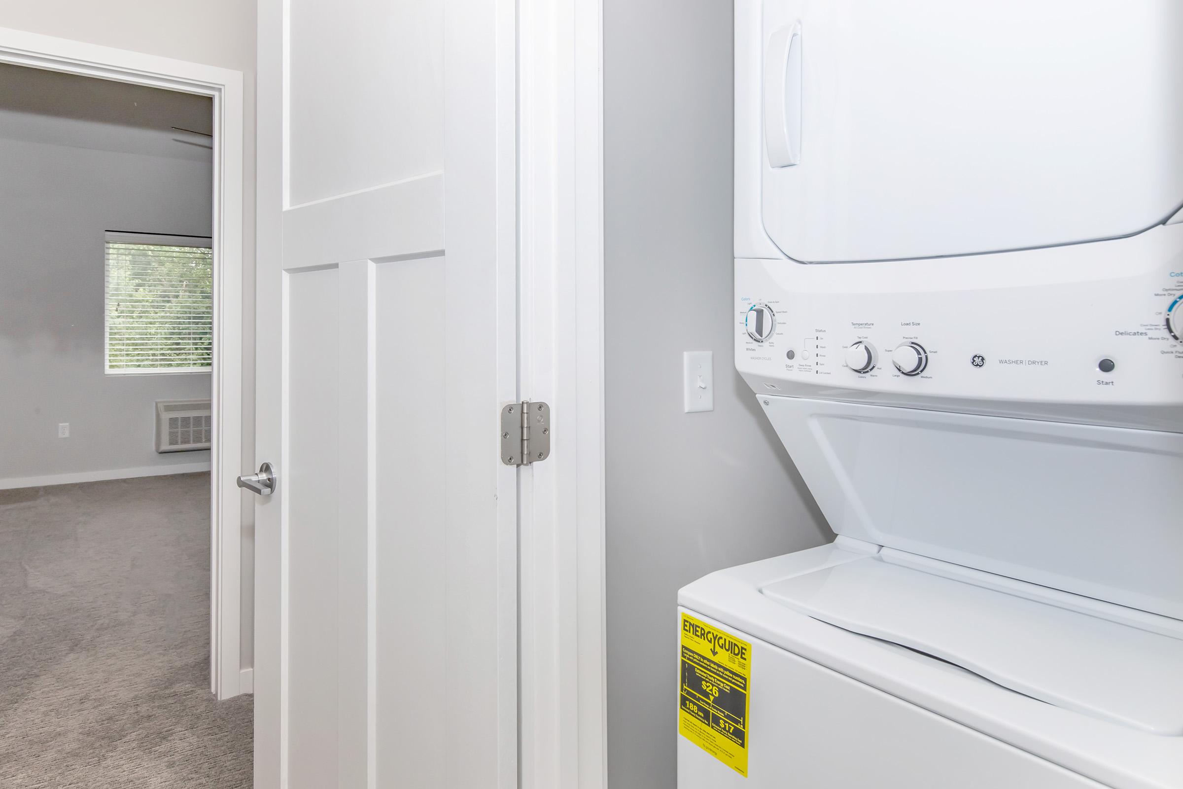 A compact laundry area featuring stacked washer and dryer units next to a partially open white door leading to a carpeted room with a window. The walls are painted in a light color, contributing to a clean, modern appearance.