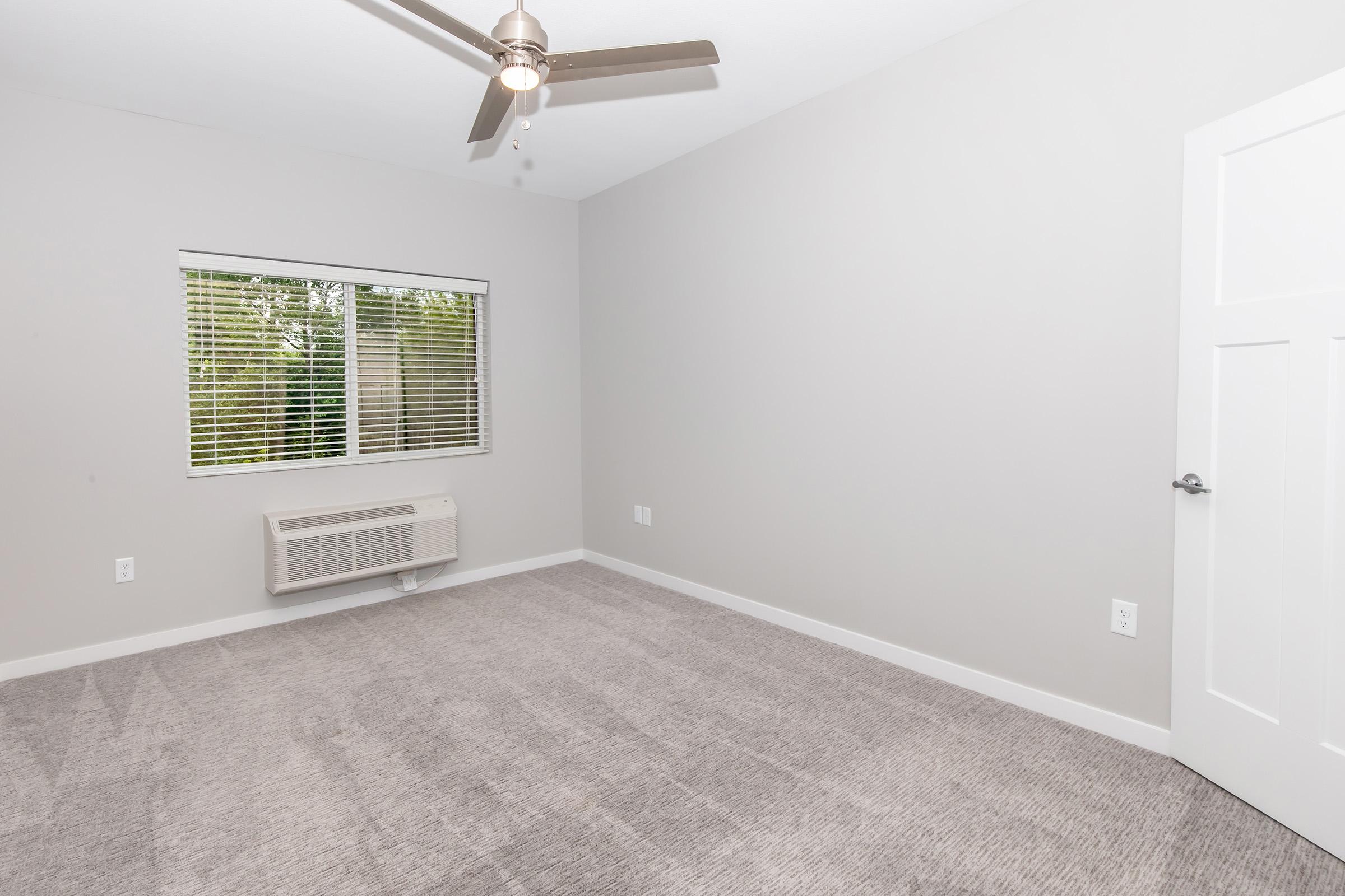 A spacious, unfurnished room with light gray walls and carpet. It features a ceiling fan, a window with horizontal blinds allowing natural light, and an air conditioning unit on the wall. A closed white door is visible, enhancing the minimalist aesthetic of the space.
