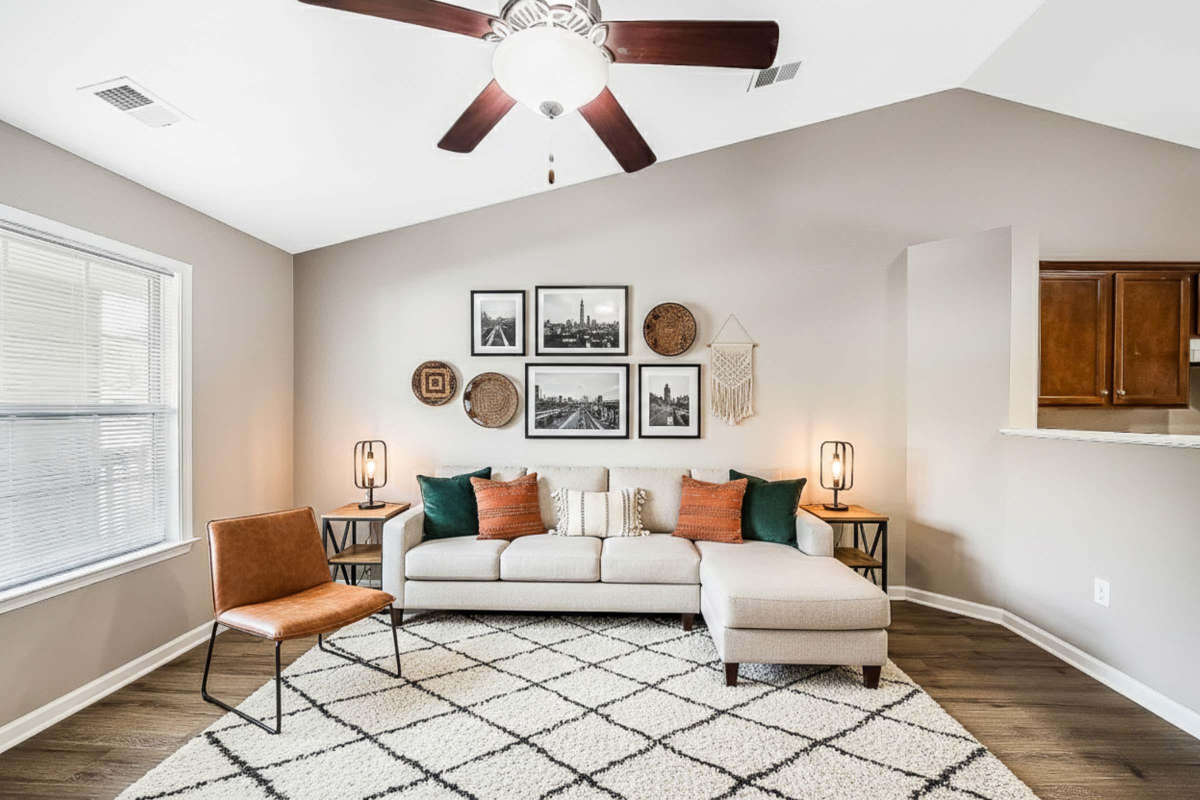 Modern living room featuring a light-colored sectional sofa with decorative pillows, a brown accent chair, and a textured area rug. The walls are adorned with framed black-and-white photographs and woven decor. A ceiling fan with light hangs above, and natural light comes through a large window.