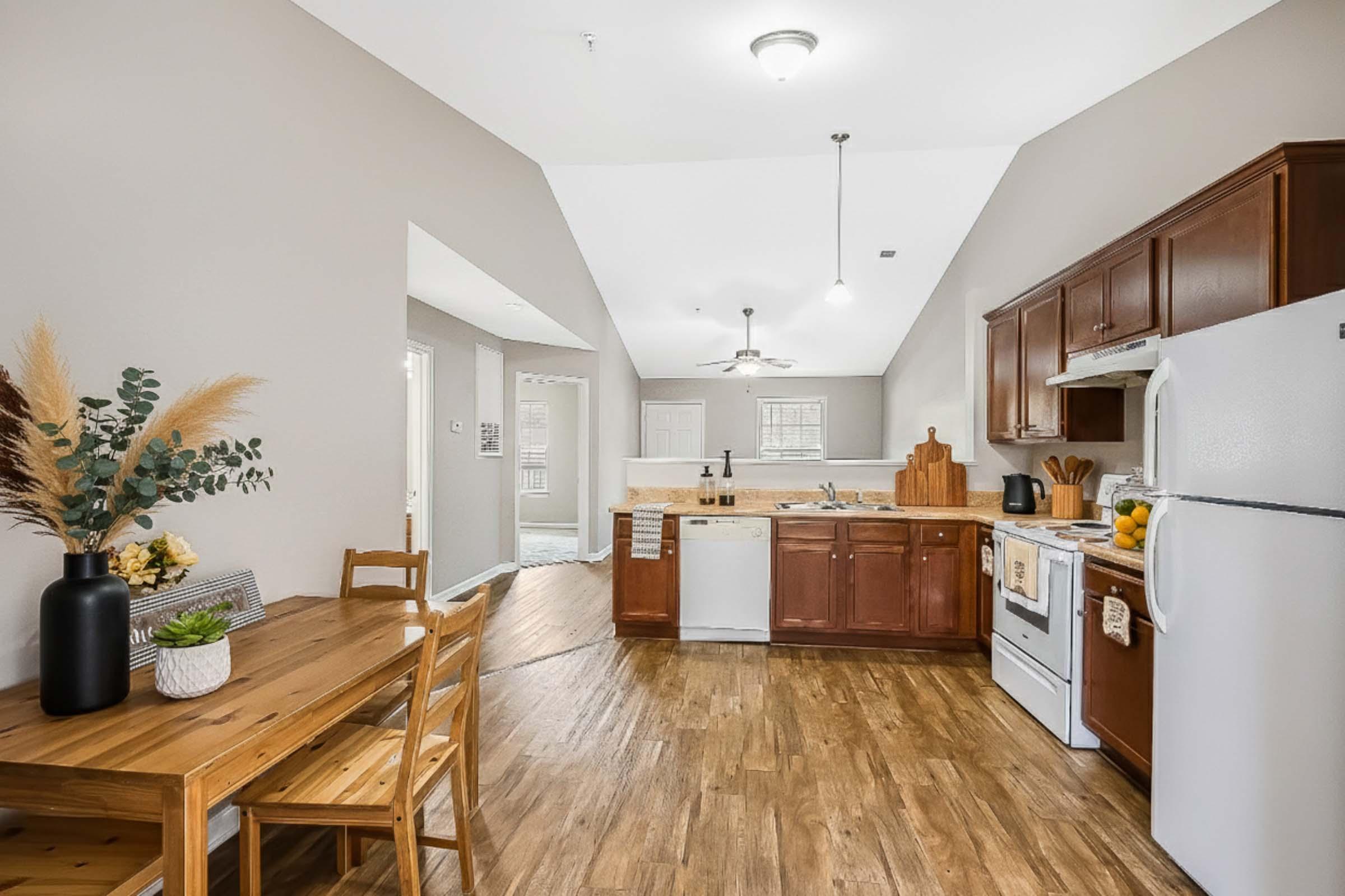 A modern kitchen with wooden cabinets and white appliances. A wooden dining table with chairs is positioned near a window. Decorative plants and a cutting board are on the counter. The space has a light-colored wall and wooden flooring, creating a warm and inviting atmosphere.