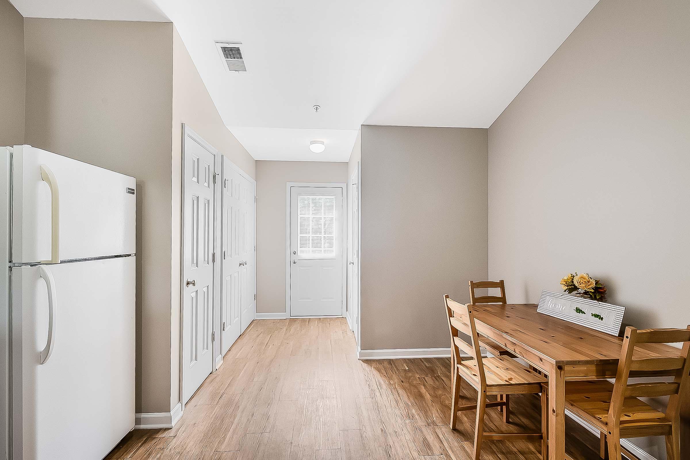 A bright and simple kitchen space featuring a white refrigerator, wooden dining table with chairs, and a small floral centerpiece. Light-colored walls and wooden flooring create a warm atmosphere. A door leads to the outside, with ample natural light streaming in through a window.