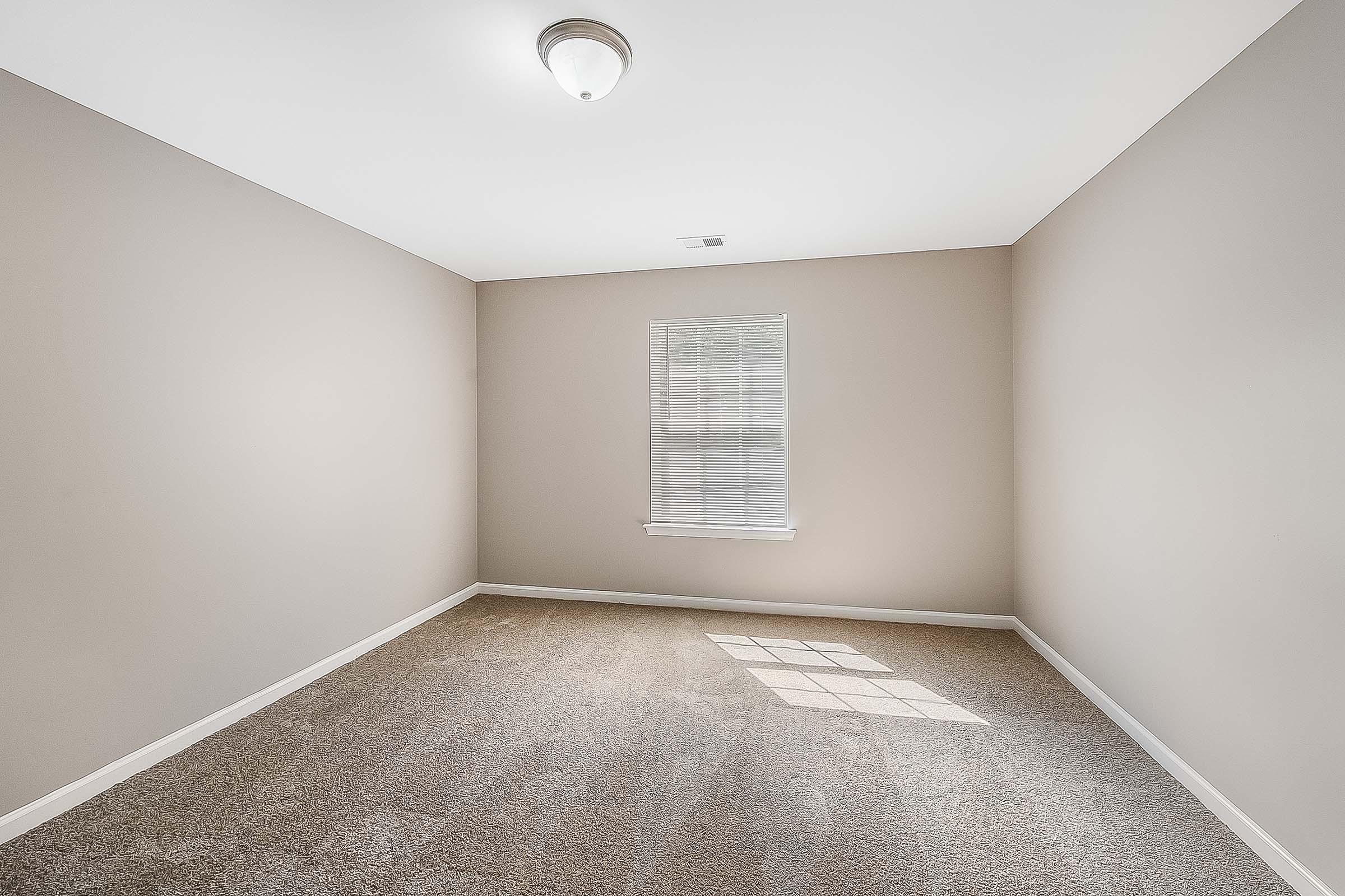 A vacant room with light beige walls and plush carpet. A small window with white blinds allows natural light to enter, casting a shadow on the floor. The ceiling features a simple light fixture, and the space appears clean and uncluttered, ready for potential furnishings.