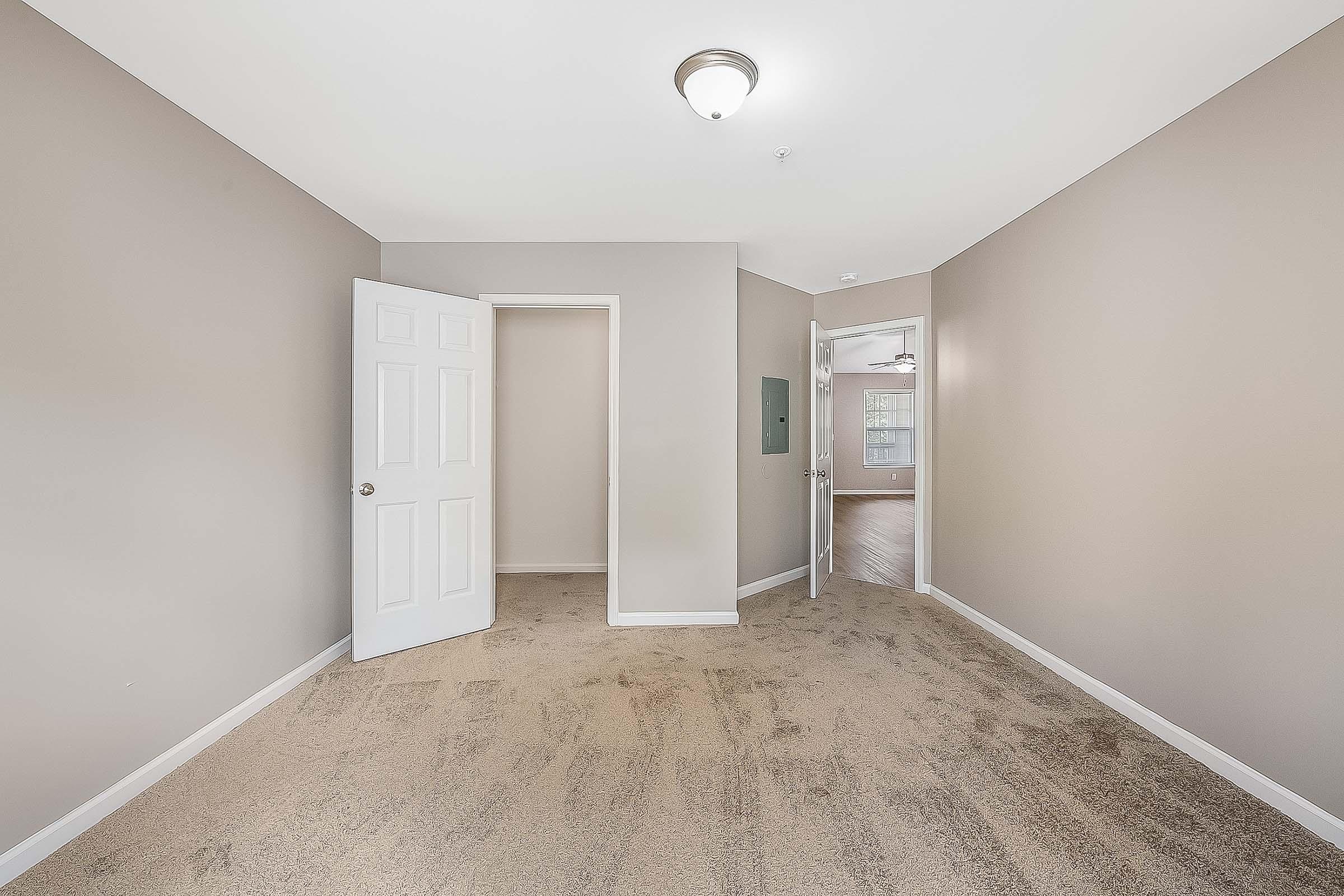 A spacious, light-colored room with beige walls and carpeted flooring. It features an open doorway leading to another area, a closet, and a ceiling light fixture. The overall atmosphere is bright and inviting, suitable for various uses such as a living area or bedroom.