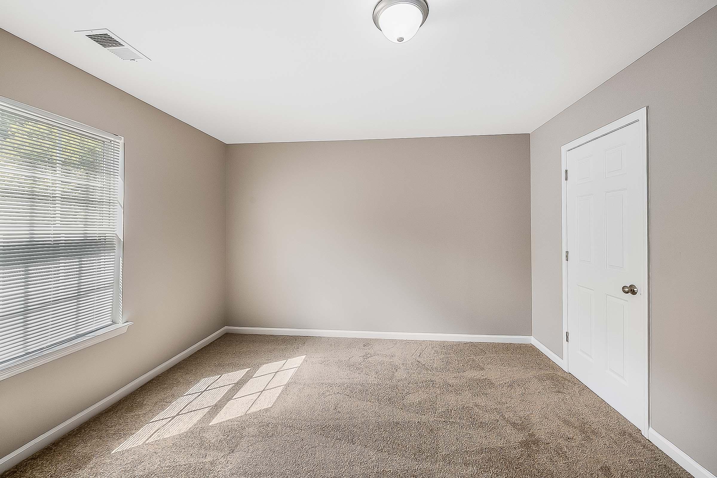 A clean, empty room with beige walls and a window adorned with blinds. Natural light filters through the window, casting shadows on the plush, carpeted floor. A white door is visible on the right side of the image, and a simple ceiling light fixture hangs above.