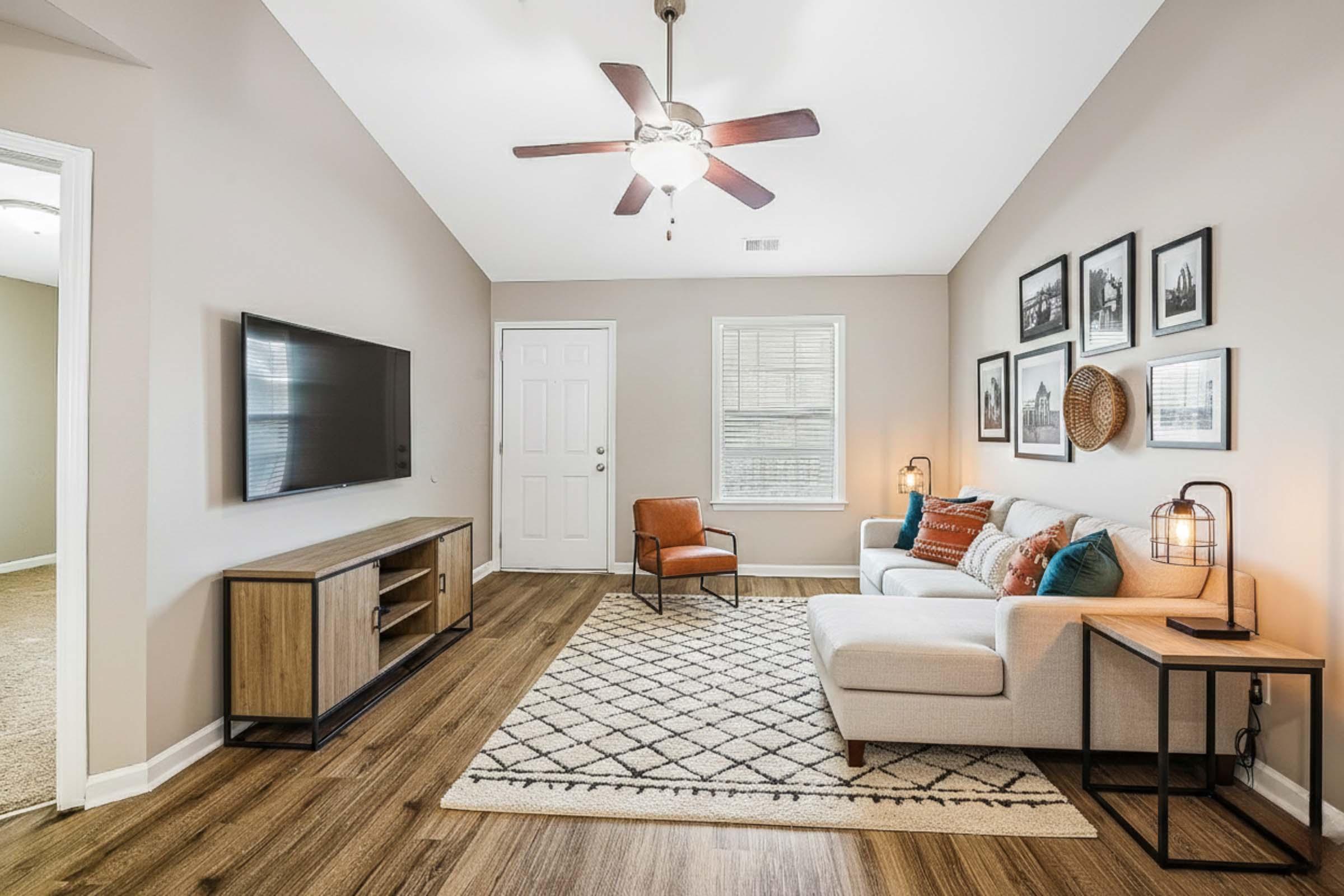 A modern living room with a light color scheme. It features a comfortable sofa with decorative pillows, a wooden coffee table, and a stylish armchair. There's a ceiling fan, a TV mounted on the wall, and framed pictures. Natural light comes in through a window beside the door, and a textured area rug adds warmth to the space.