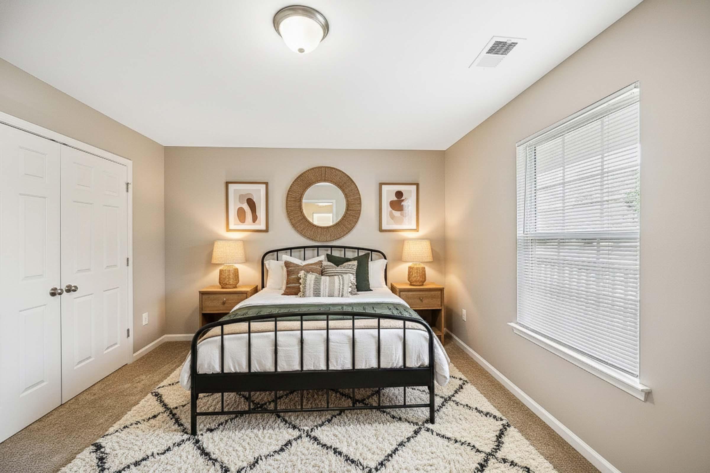 A cozy bedroom featuring a black metal bed frame with white bedding, flanked by two wooden nightstands with lamps. Above the bed are two framed artworks. A round mirror hangs on the wall, and the room has beige walls and carpet. It includes a large window with blinds, allowing natural light to enter.