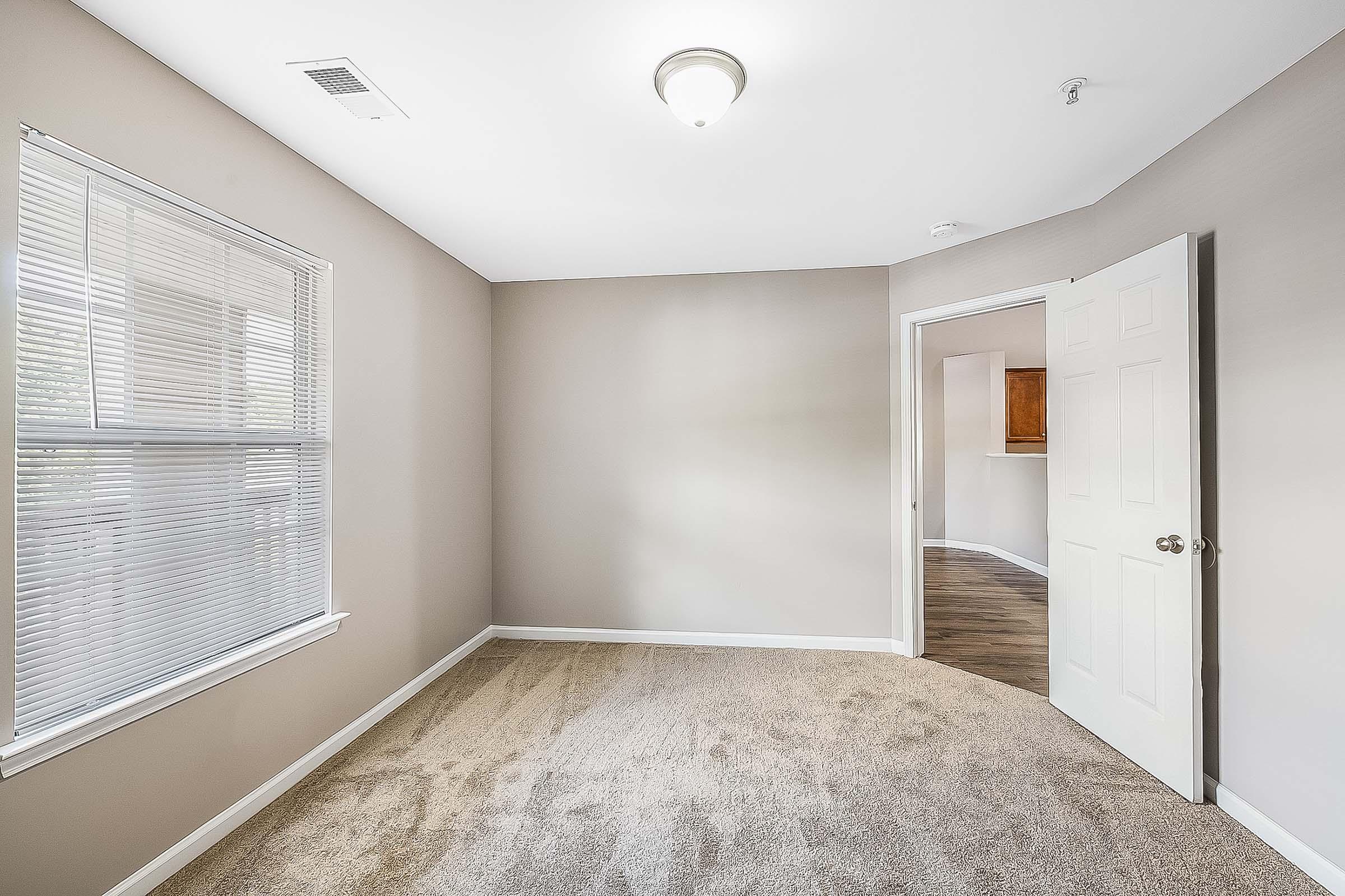 Empty room with beige walls and carpet, featuring a window with blinds, a ceiling light, and an open doorway leading to another space. The room has a clean and simple design, suitable for various uses or decorating options.