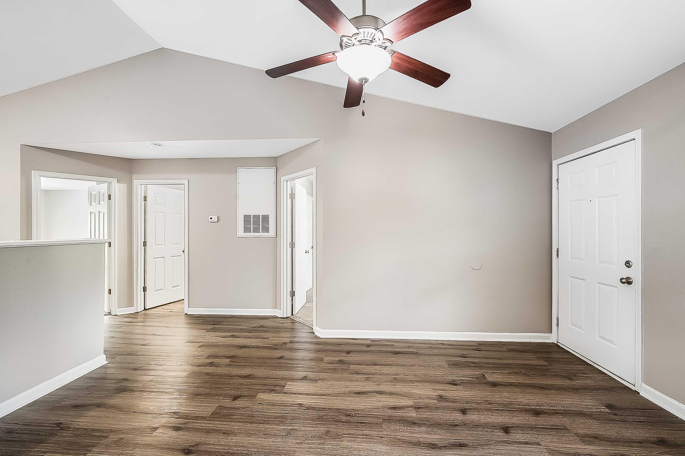 A spacious interior room with wooden flooring, featuring a ceiling fan with light. Light-colored walls and a high ceiling create an airy atmosphere. Doorways lead to adjacent rooms, while a front door is visible on the right, enhancing accessibility and flow within the space.