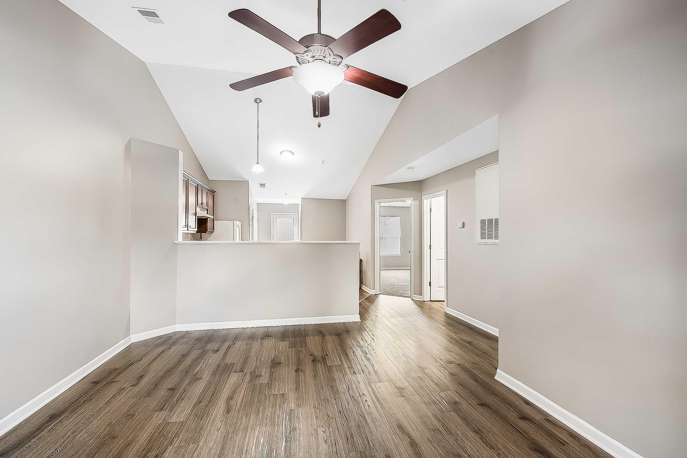 Interior view of a spacious, well-lit room featuring a ceiling fan, wood-style flooring, and neutral-colored walls. The layout includes an open area leading to a kitchen with wooden cabinets and a doorway to another room. Natural light enters through an overhead light fixture and additional doorways.