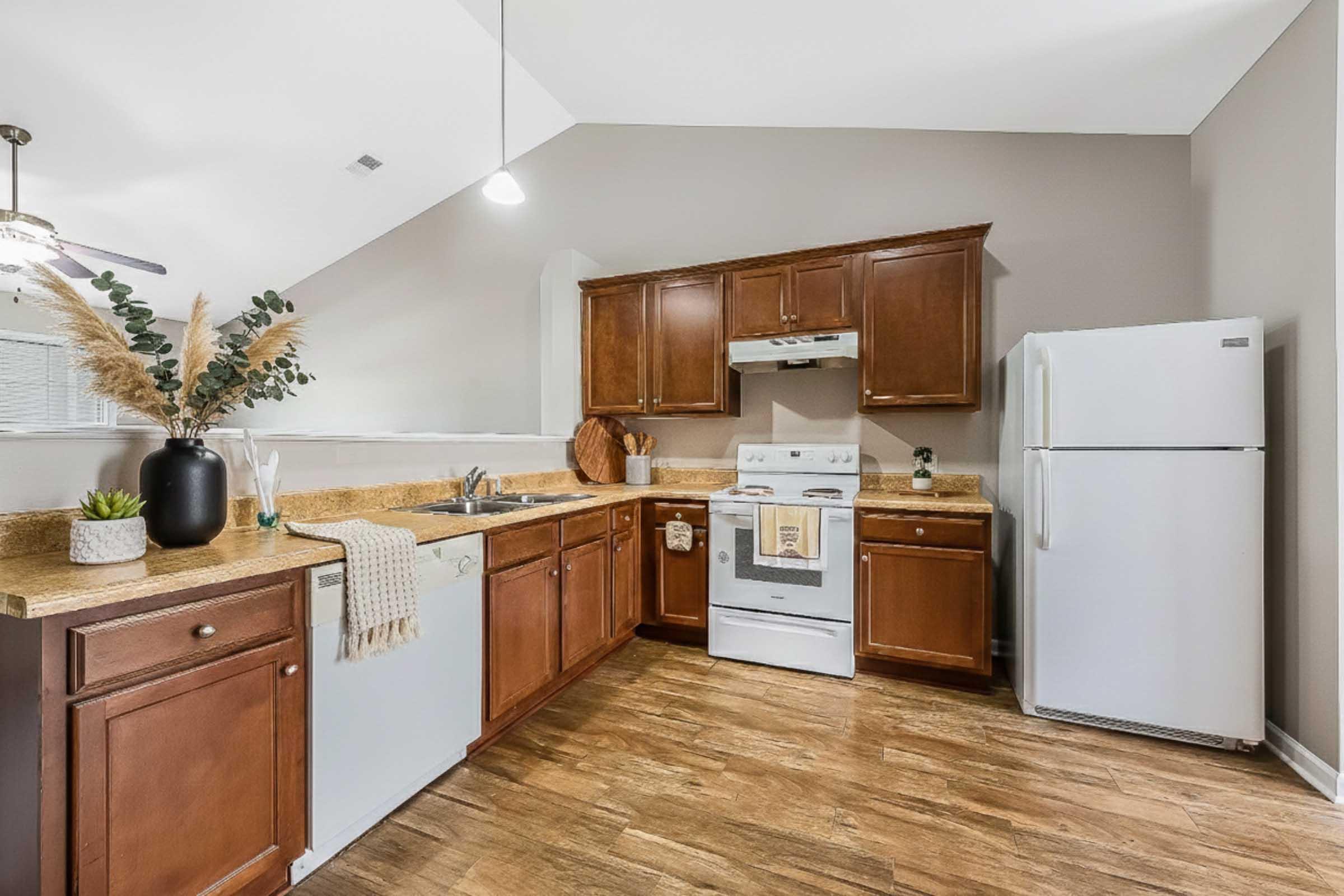 A modern kitchen featuring wooden cabinetry, a white refrigerator, a stove with an overhead exhaust, and a dishwasher. The countertops are light-colored with decorative elements like a plant and a white dish towel. Natural light enters through a nearby window, enhancing the spacious feel of the room.