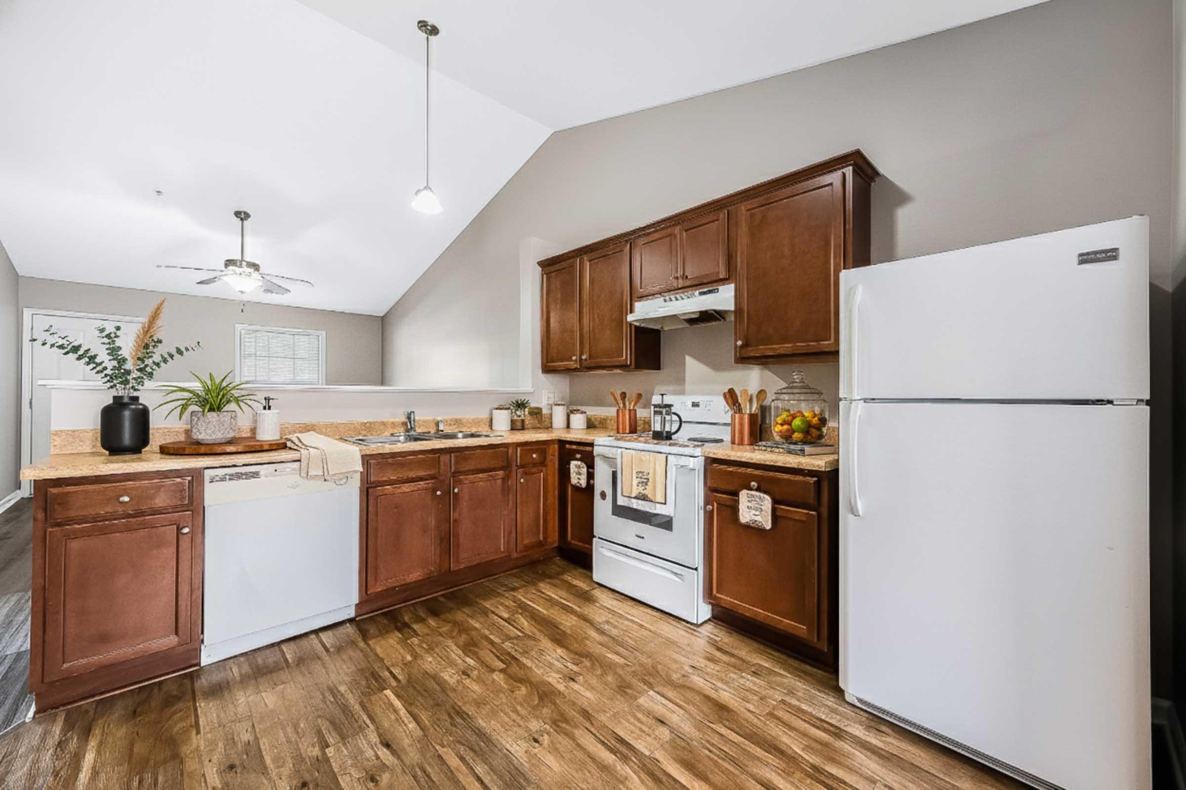 Modern kitchen featuring wooden cabinets, a white refrigerator, stove, and dishwasher. The countertop is adorned with kitchen utensils and a fruit bowl. Natural light enters through a window, highlighting the warm wood flooring and neutral wall colors. A ceiling fan adds functionality to the space.