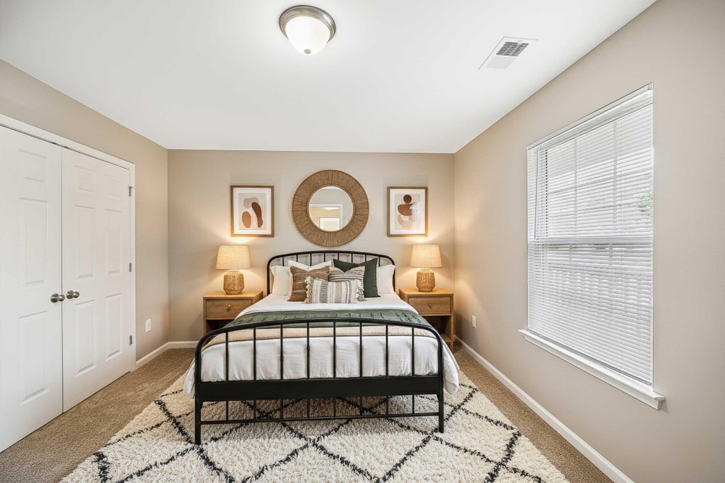 A cozy bedroom with a stylish black metal bed frame, adorned with crisp white bedding and decorative pillows. Two bedside tables with lamps flank the bed, and abstract art is displayed on the walls. A round mirror reflects light, and a plush area rug adds warmth to the space. Natural light streams in through a window.