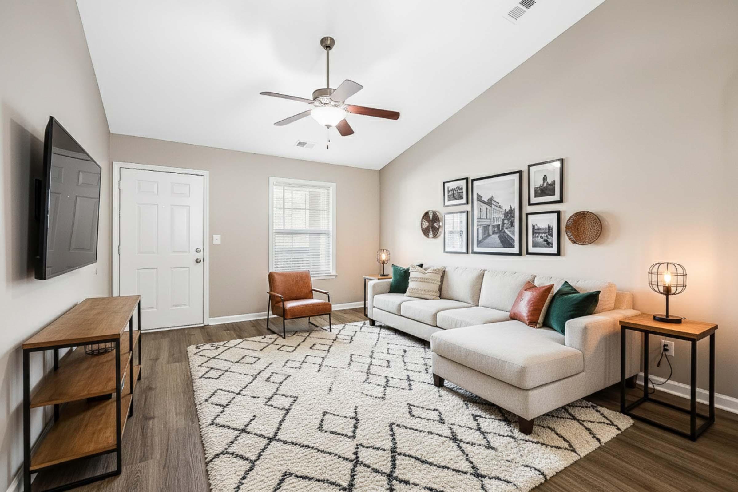 A modern living room featuring a beige sectional sofa with colorful cushions, a wooden armchair, a television mounted on the wall, and a decorative area rug with geometric patterns. There are framed black-and-white photographs and wall decor, and a ceiling fan adds to the ambiance. Natural light comes through the window.