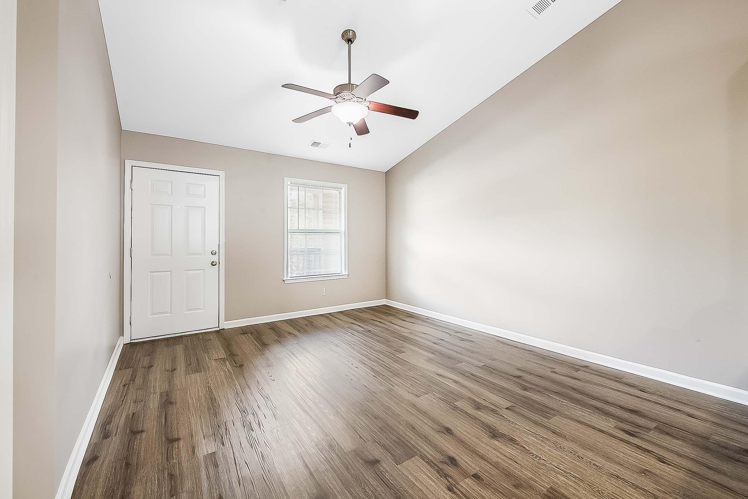 A well-lit, empty room with light brown walls and a ceiling fan. The floor is made of wood-like material. There's a white door on the left and a window on the right, allowing natural light to enter the space. The room has a high ceiling, creating an open and airy feel.