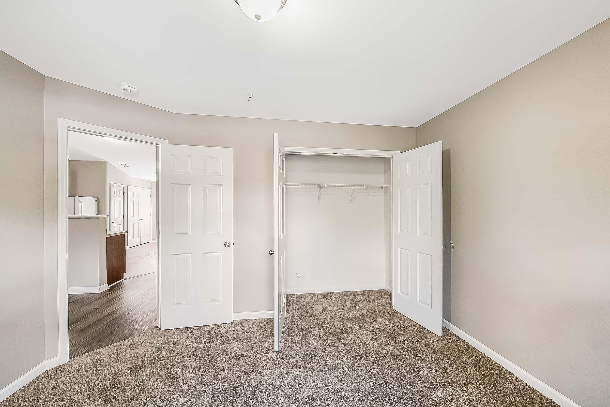 Interior view of a cozy room with light beige walls and plush carpet. Two white double doors are open to reveal a spacious closet with hanging space. A doorway leads to another part of the house, showcasing a glimpse of the living area in the background. Natural light illuminates the space.