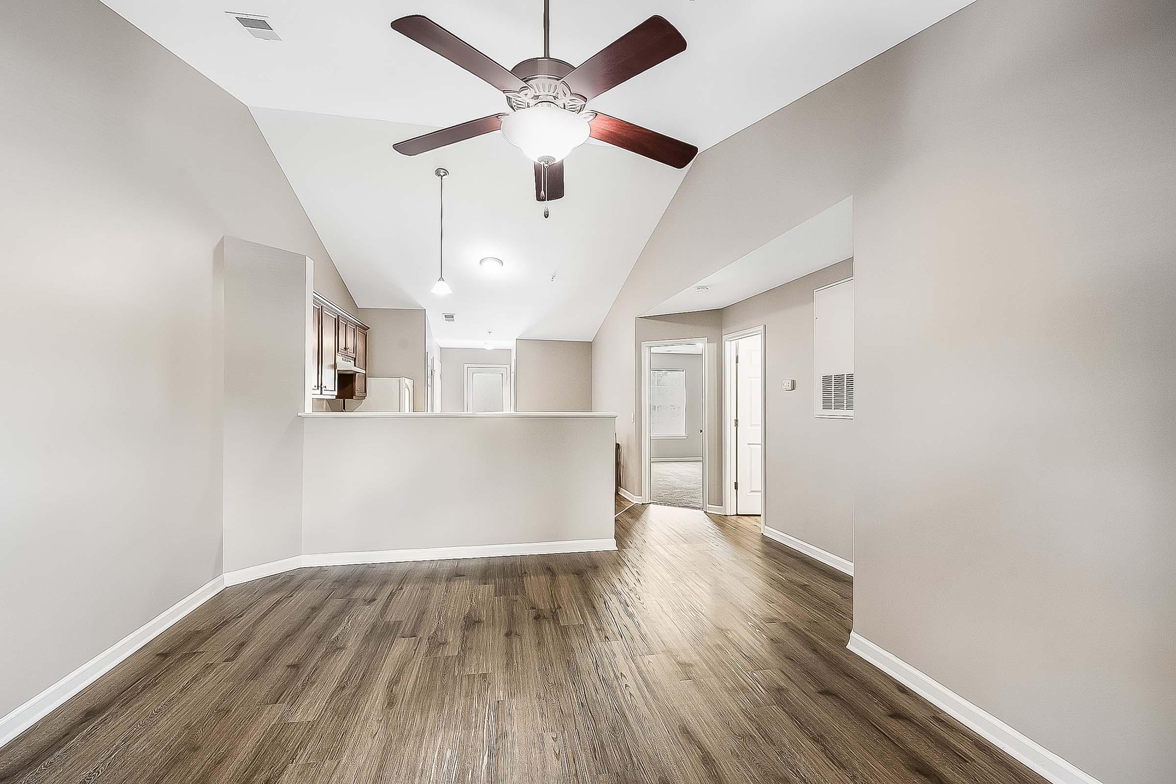 Spacious interior view of a modern living area, featuring hardwood flooring, a ceiling fan, and soft gray walls. A partially visible kitchen area is on the left, with an open layout leading to a bright hallway and a door at the end, suggesting access to additional rooms. Natural light enhances the inviting atmosphere.