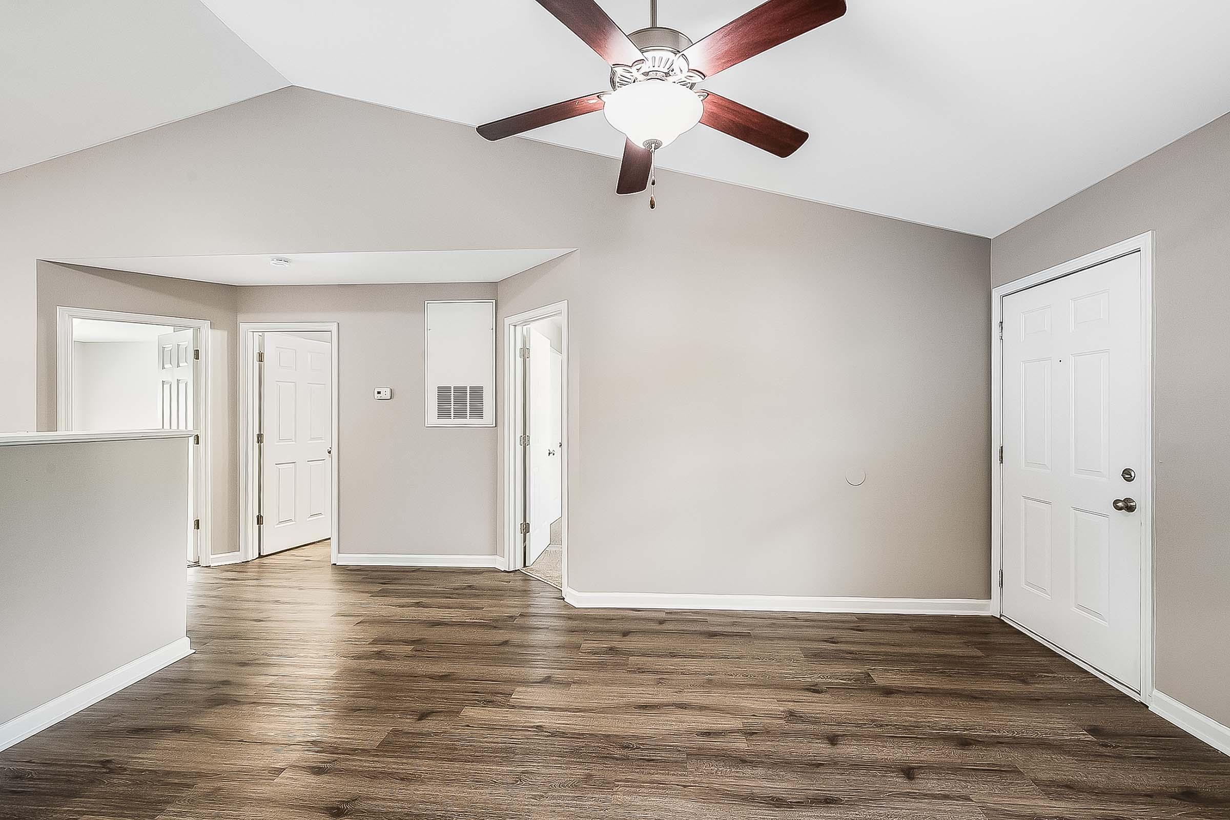 Interior view of a spacious room featuring a ceiling fan, wooden flooring, and light beige walls. There are doorways leading to other areas, including a white door on the right. The room has a bright and open feel with natural light coming from unseen windows.