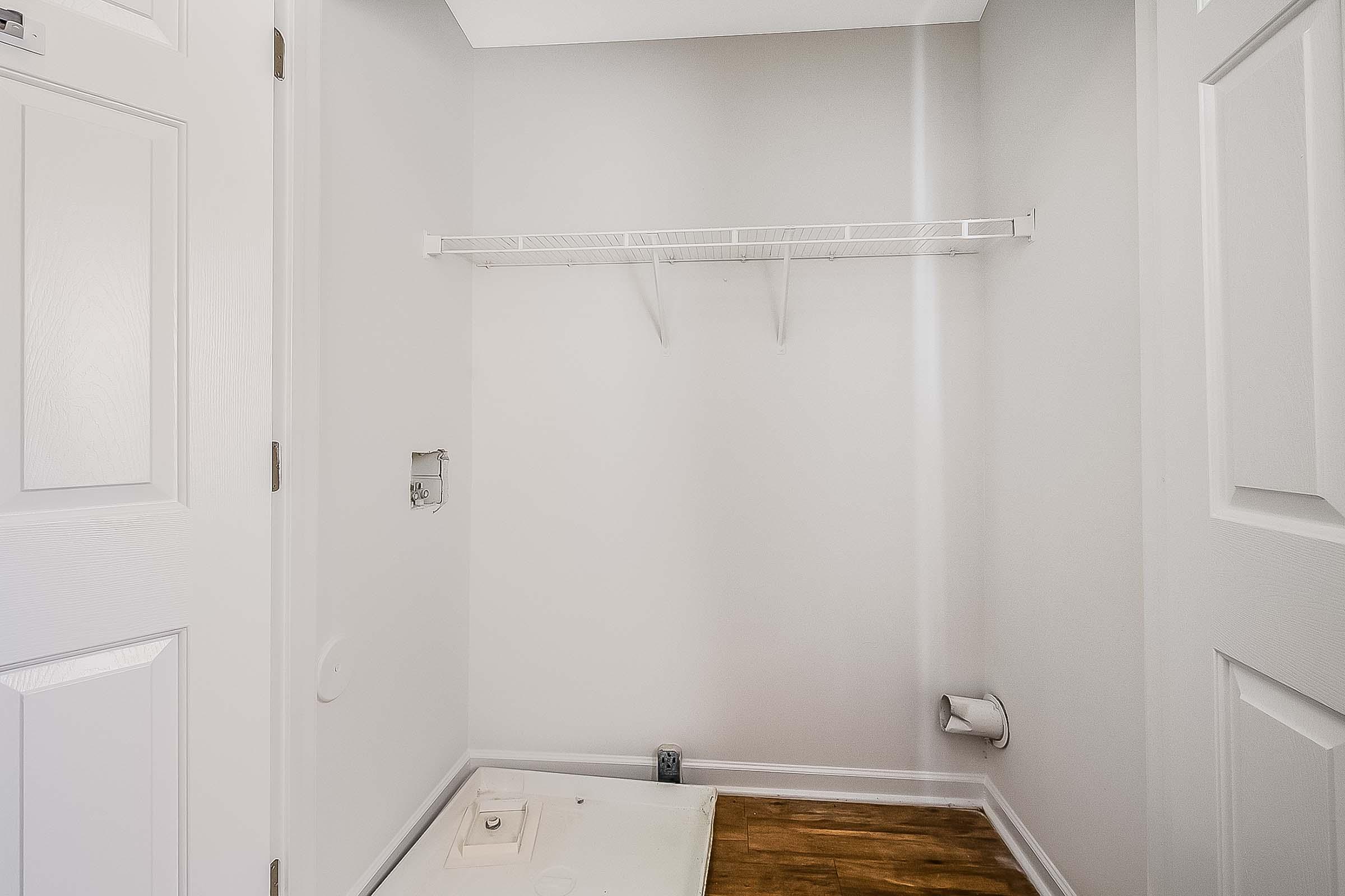Empty laundry room with light gray walls, featuring a white shelf mounted on the wall. There is a small opening in the wall for plumbing, and a space on the floor where a washer or dryer could be placed. The room has a wooden floor and a door visible in the frame.