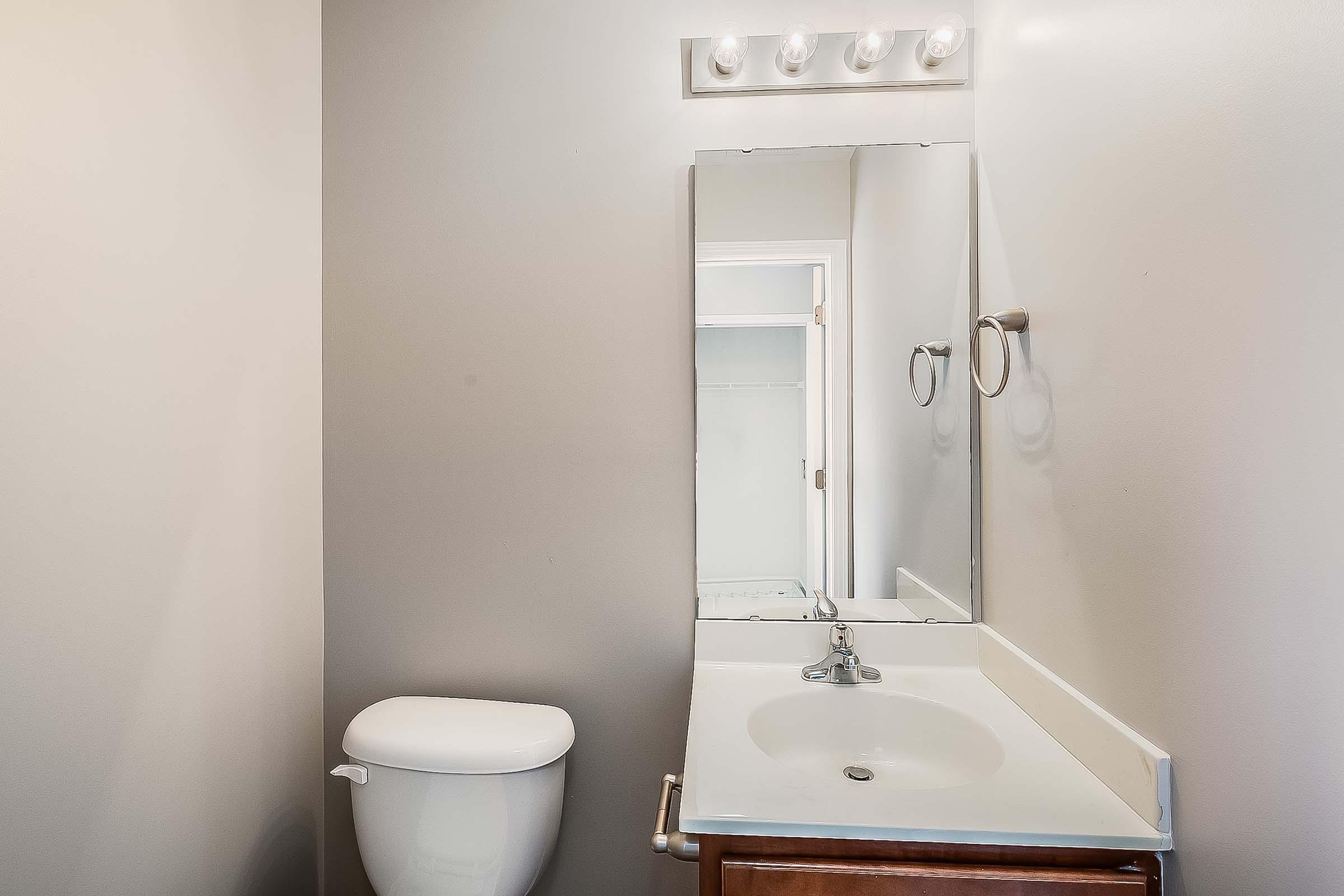 A clean and simple bathroom featuring a white toilet, a small sink with a faucet, and a large mirror. Above the mirror, there are five light fixtures. The walls are painted in a neutral tone, creating a spacious feel. A doorway in the background leads to another room.