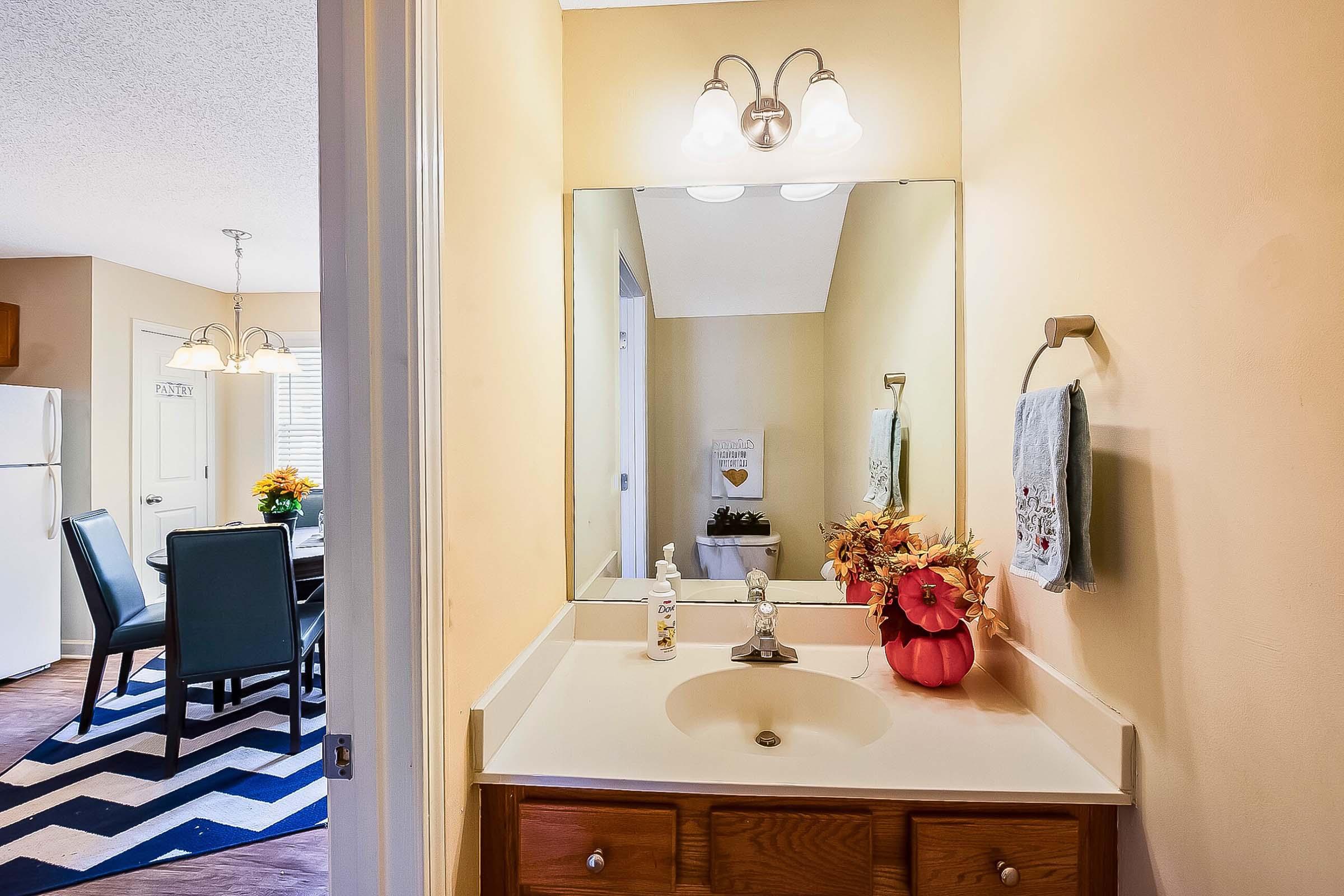 A small bathroom with a vanity sink, mirror, and decorative items. The wall is painted a light yellow, and a towel hangs on the side. In the background, a dining area with a zigzag rug, dining table, and chairs is visible. A bright arrangement of flowers adds color to the scene.