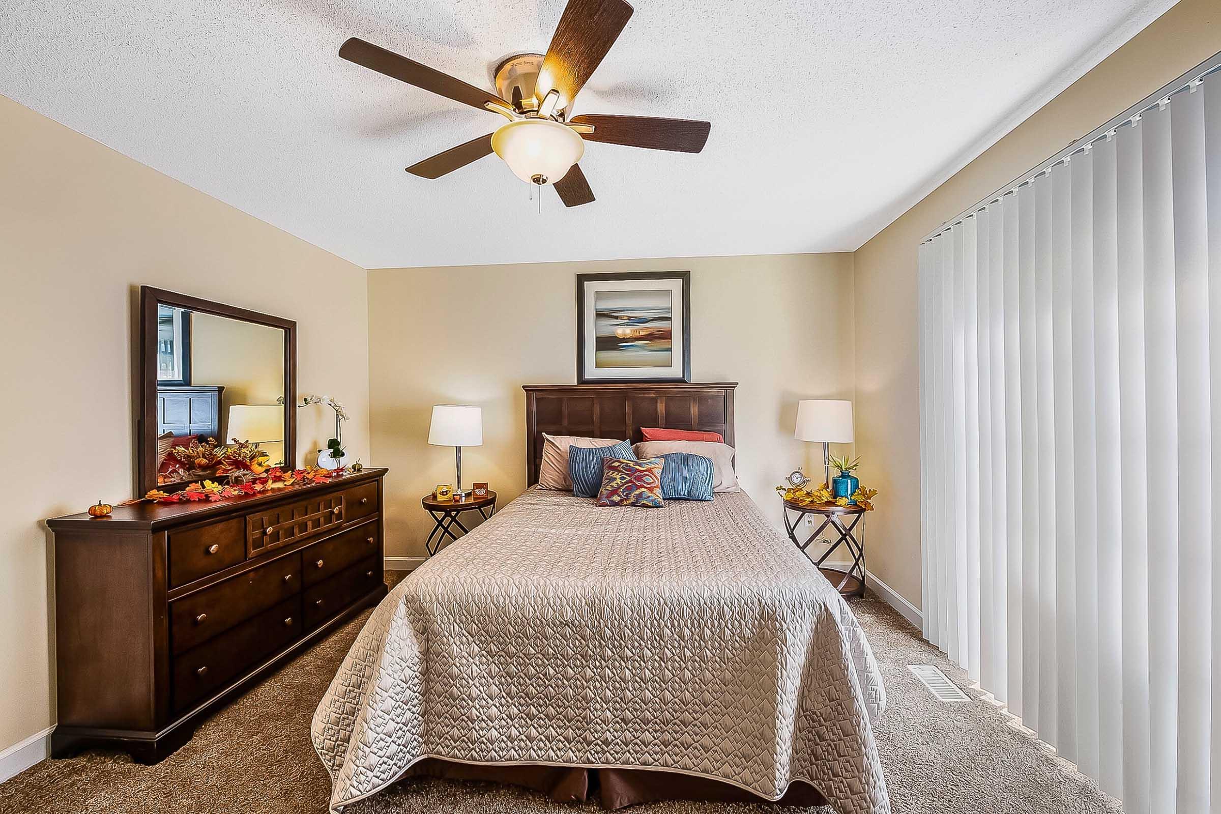 A cozy bedroom featuring a queen-sized bed with a neutral quilt, two bedside tables with lamps, and a dresser with decorative items. A large mirror reflects the room, and there are autumn-themed decorations on the dresser. The walls are painted a warm beige, and a ceiling fan hangs above.