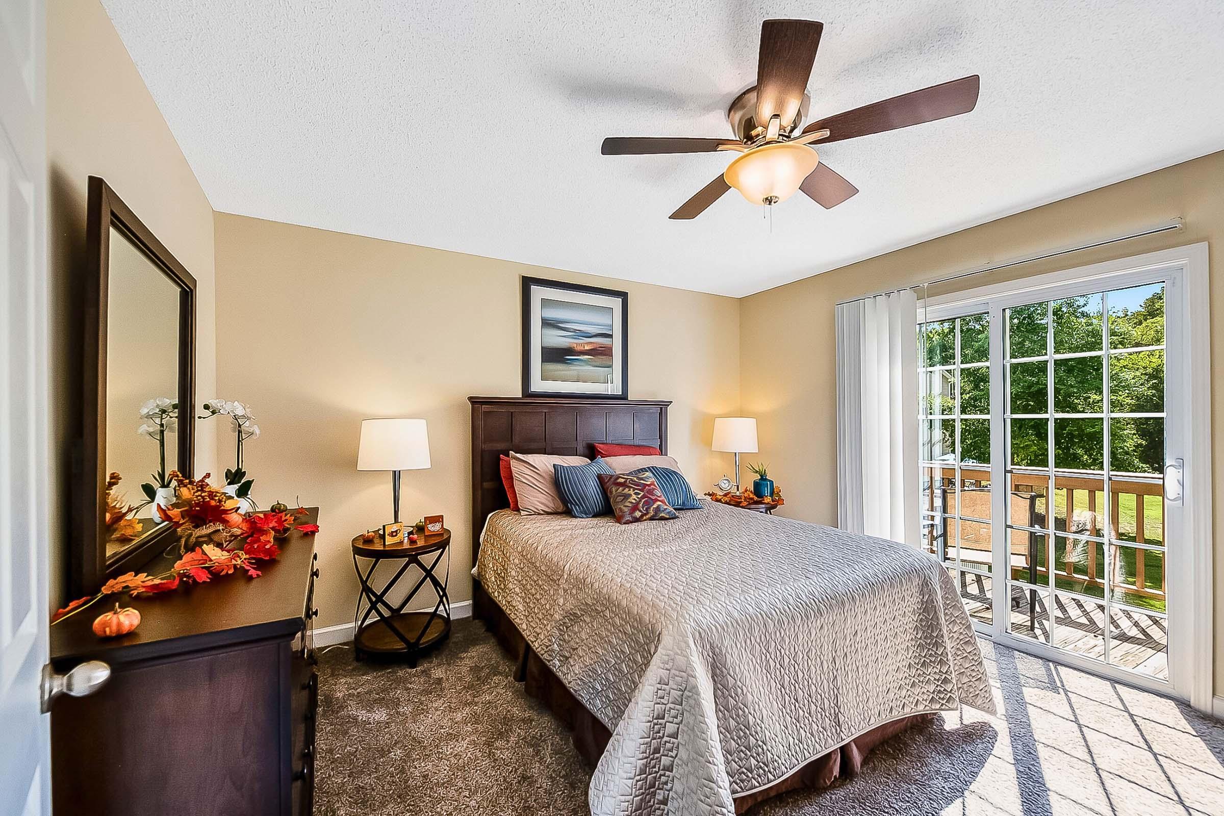Cozy bedroom featuring a queen-sized bed with decorative pillows, two bedside lamps, a large mirror, and a wooden dresser. A ceiling fan and natural light from sliding glass doors lead to an outdoor deck. Decorative fall-themed accents are placed on the bedside table. Warm, neutral wall colors enhance the inviting atmosphere.