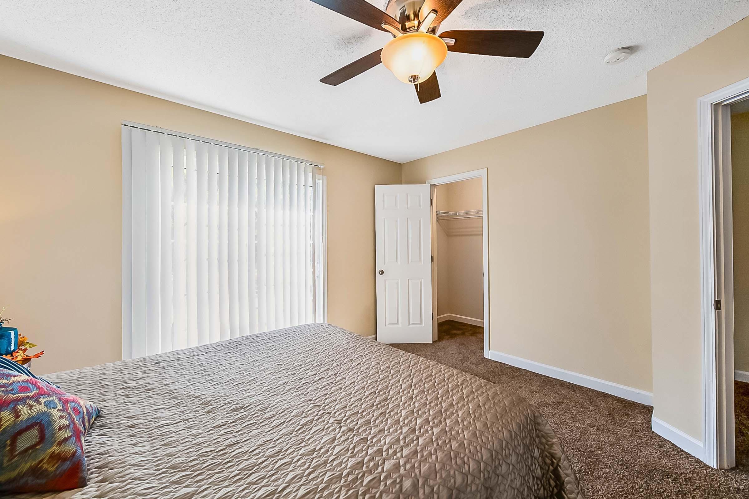 A cozy bedroom featuring a queen-sized bed with a textured brown quilt, a decorative pillow, and a ceiling fan. The room has light beige walls, a large window with vertical blinds, and a door leading to a closet. Warm natural light fills the space, creating a welcoming ambiance.