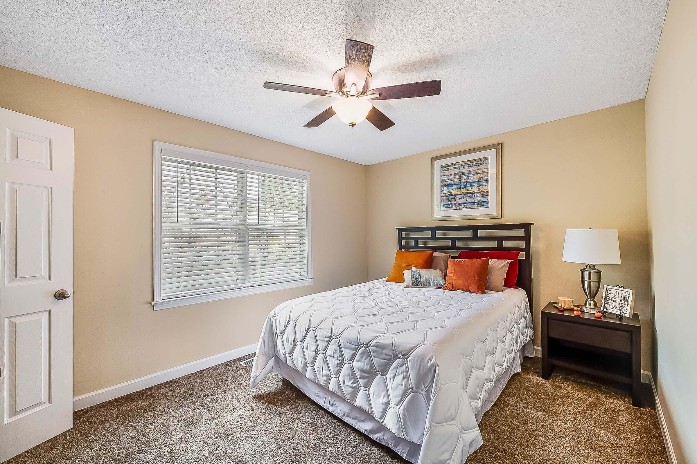 A cozy bedroom featuring a queen-sized bed with a white quilt and colorful pillows, a nightstand with a lamp and framed photos, and a ceiling fan. A window with blinds allows natural light in, and the walls are painted a warm beige tone with a decorative piece of art above the bed.