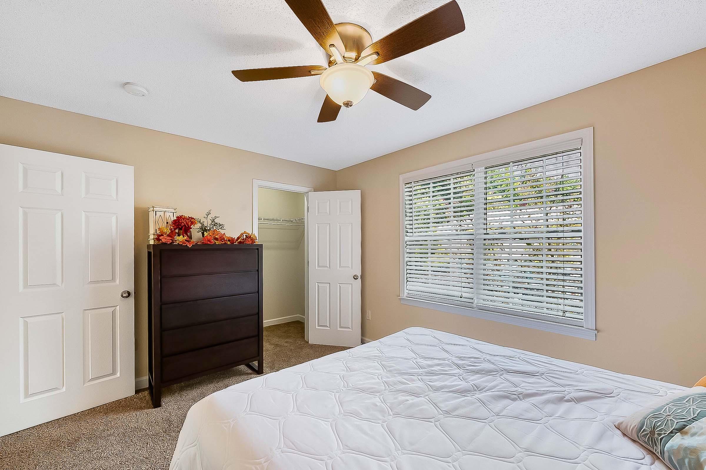 A cozy bedroom featuring a white bed with a quilted blanket, a dark wooden dresser, and a ceiling fan. The room has beige walls, a window with white blinds letting in natural light, and two closed doors. Decorative autumn leaves are placed on the dresser for a seasonal touch.