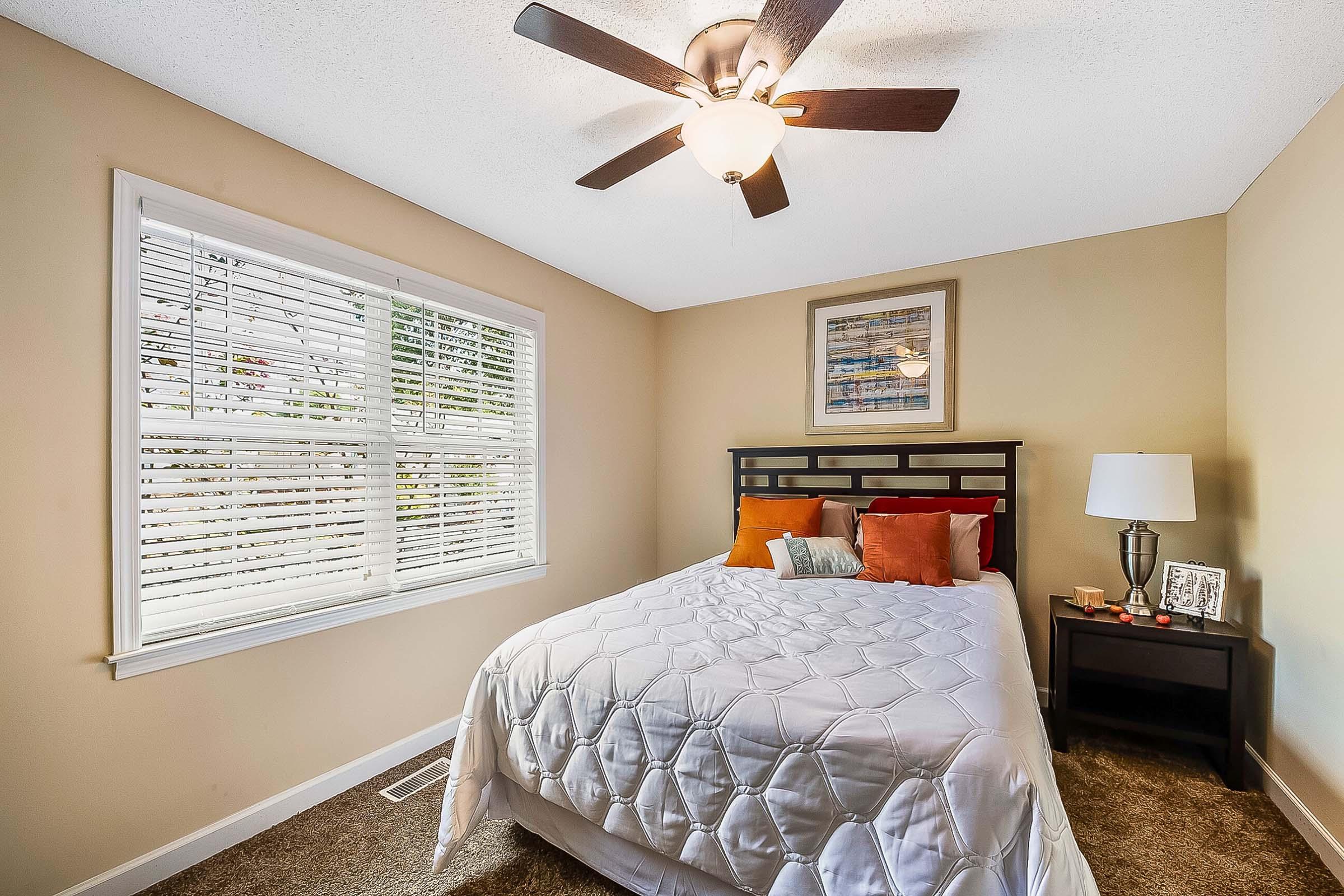Cozy bedroom featuring a queen bed with white bedding and orange accents, a window with blinds allowing natural light, a nightstand with a lamp and decorative items, and a ceiling fan. The walls are painted a soft beige, creating a warm and inviting atmosphere.