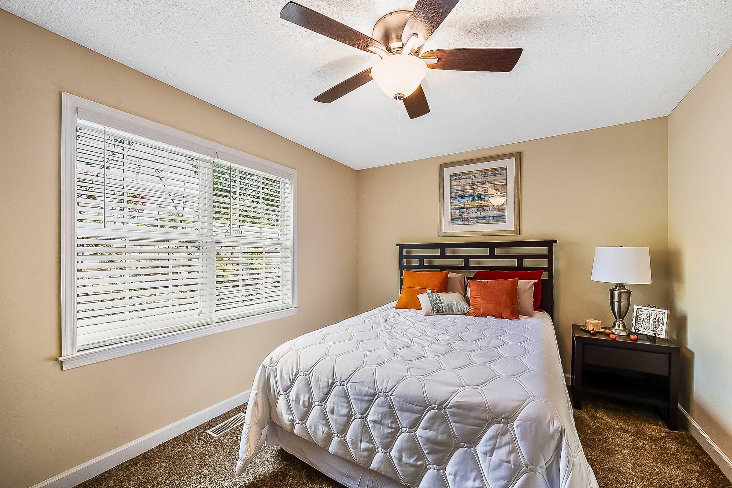 Cozy bedroom featuring a queen-sized bed with white bedding and orange accent pillows, a bedside table with a lamp and picture frame, a window with white blinds allowing natural light, and a ceiling fan overhead. Neutral walls and carpet create a warm atmosphere.