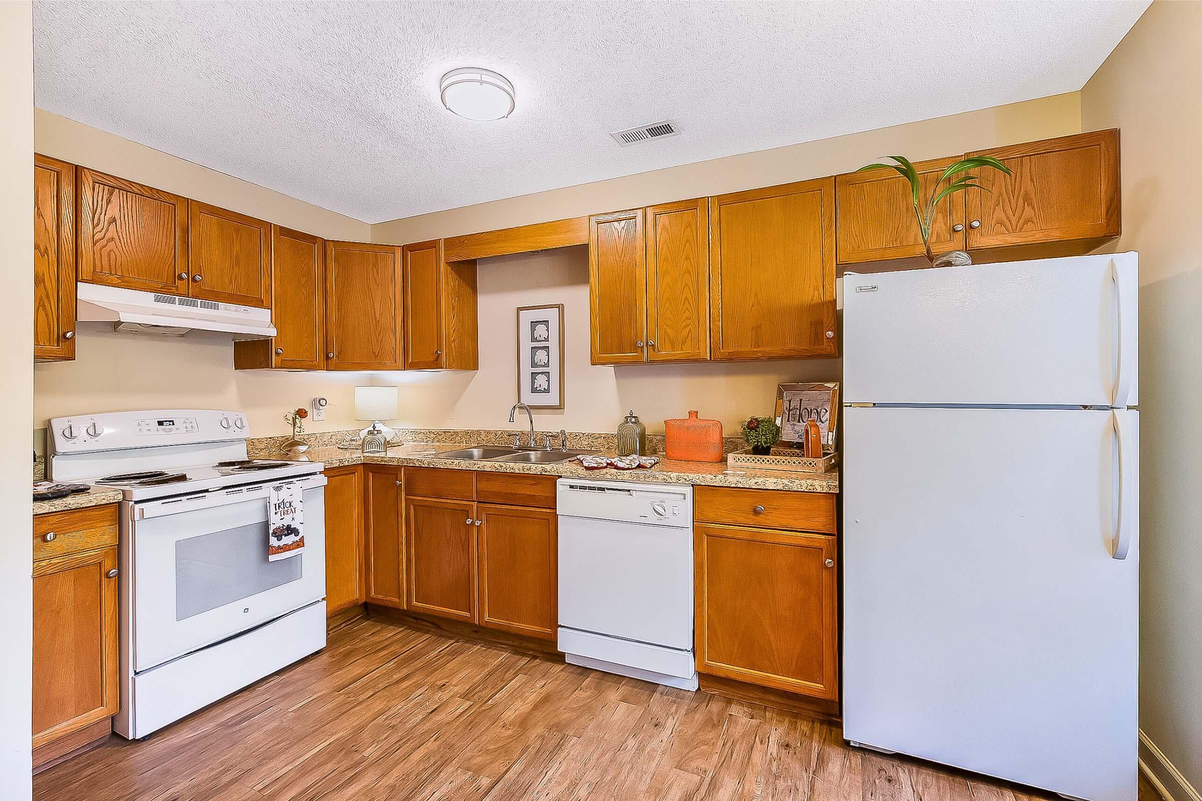 A cozy kitchen with wooden cabinets, a white stove, and a dishwasher. The countertop features a sink and decorative items, while a white refrigerator stands in the corner. The warm-toned flooring enhances the inviting atmosphere of the space.