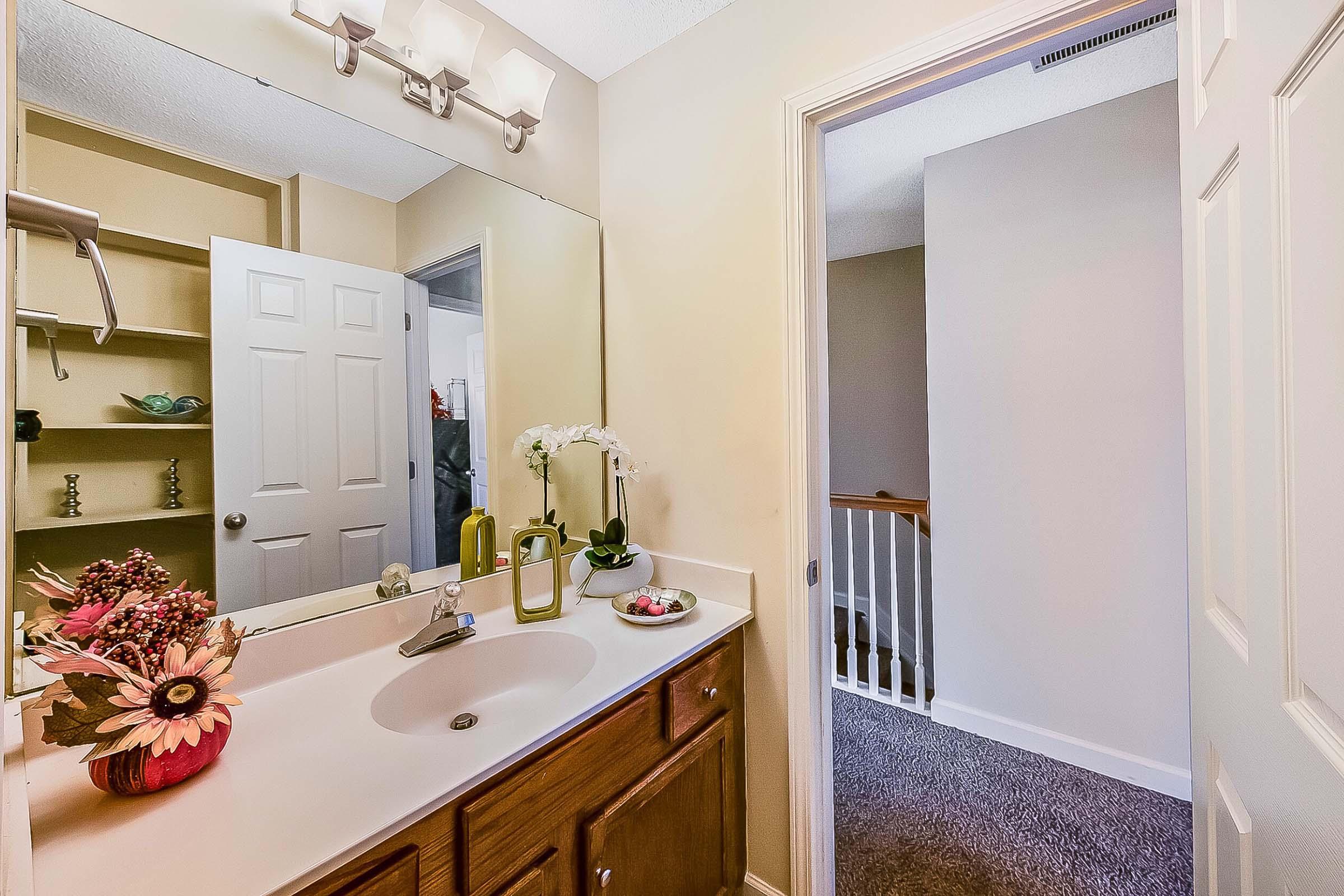 A well-lit bathroom featuring a double sink vanity with a large mirror above, decorative flowers, and a small plant. A door leads to a hallway with carpeted flooring visible in the background, while a closet with shelves is partially open on the left side.
