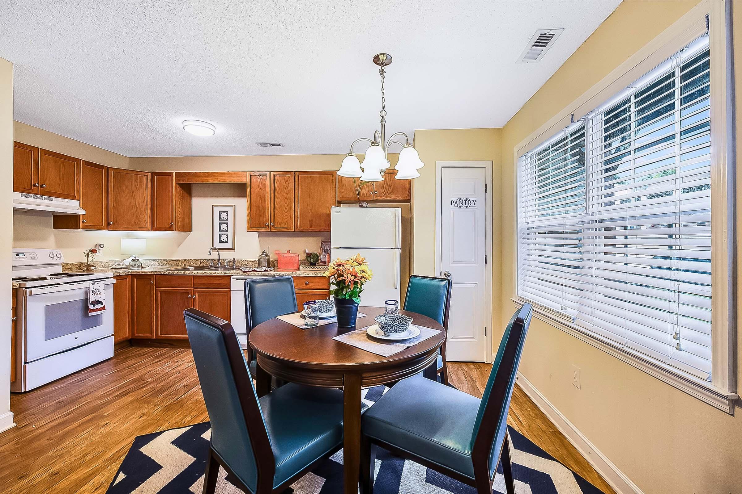 A cozy dining area featuring a round wooden table with blue chairs, set with a plate and tea cup. In the background, there is a kitchen with wooden cabinets, a white refrigerator, and a stove. Natural light streams through a window with blinds, creating a warm atmosphere. The walls are painted a soft yellow.