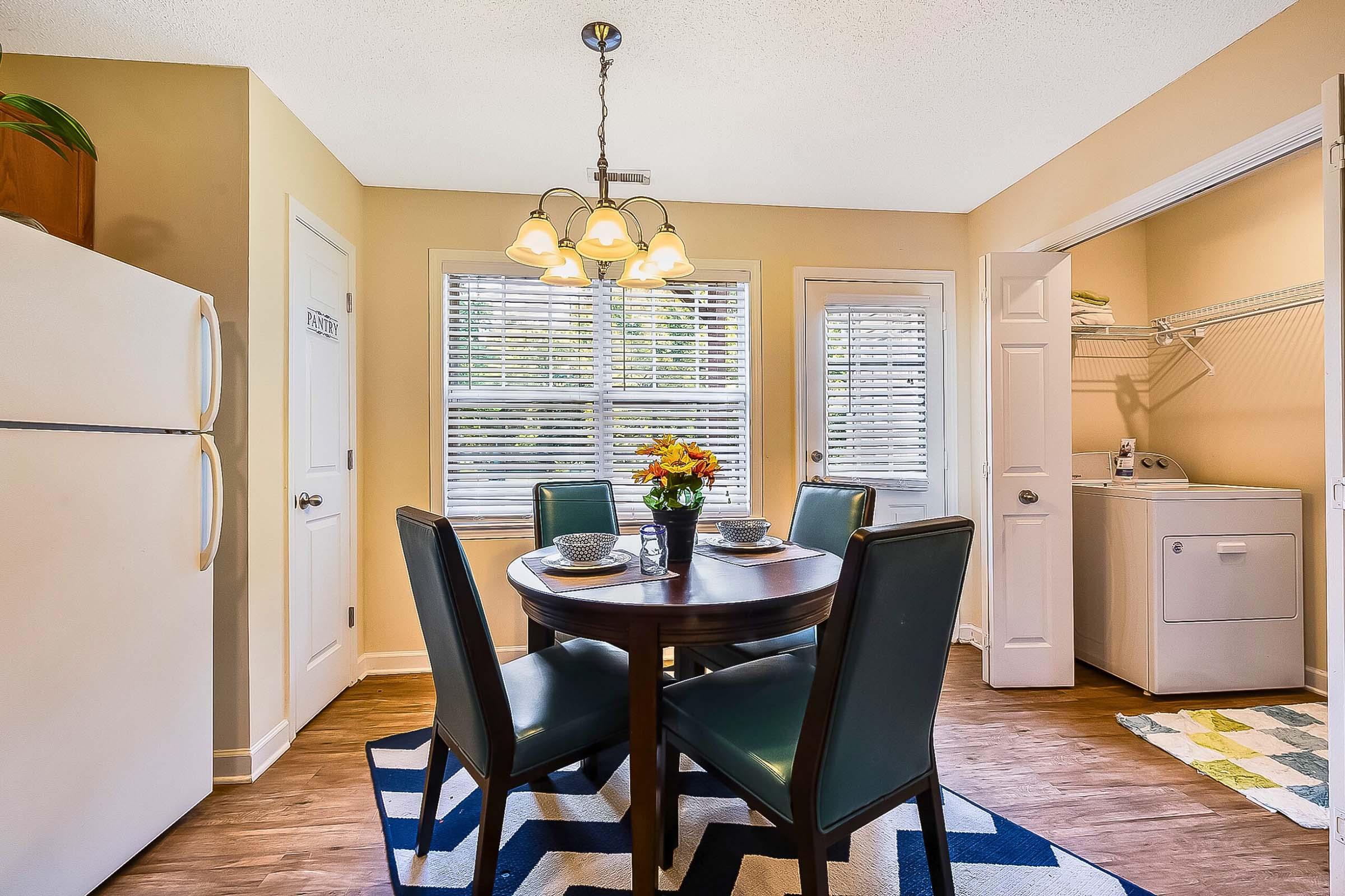 A cozy dining area featuring a round wooden table set with plates and utensils, surrounded by four green chairs. The space includes a refrigerator and a doorway leading to a laundry area. Natural light brightens the room, highlighting the simple decor and a vase of flowers at the table.