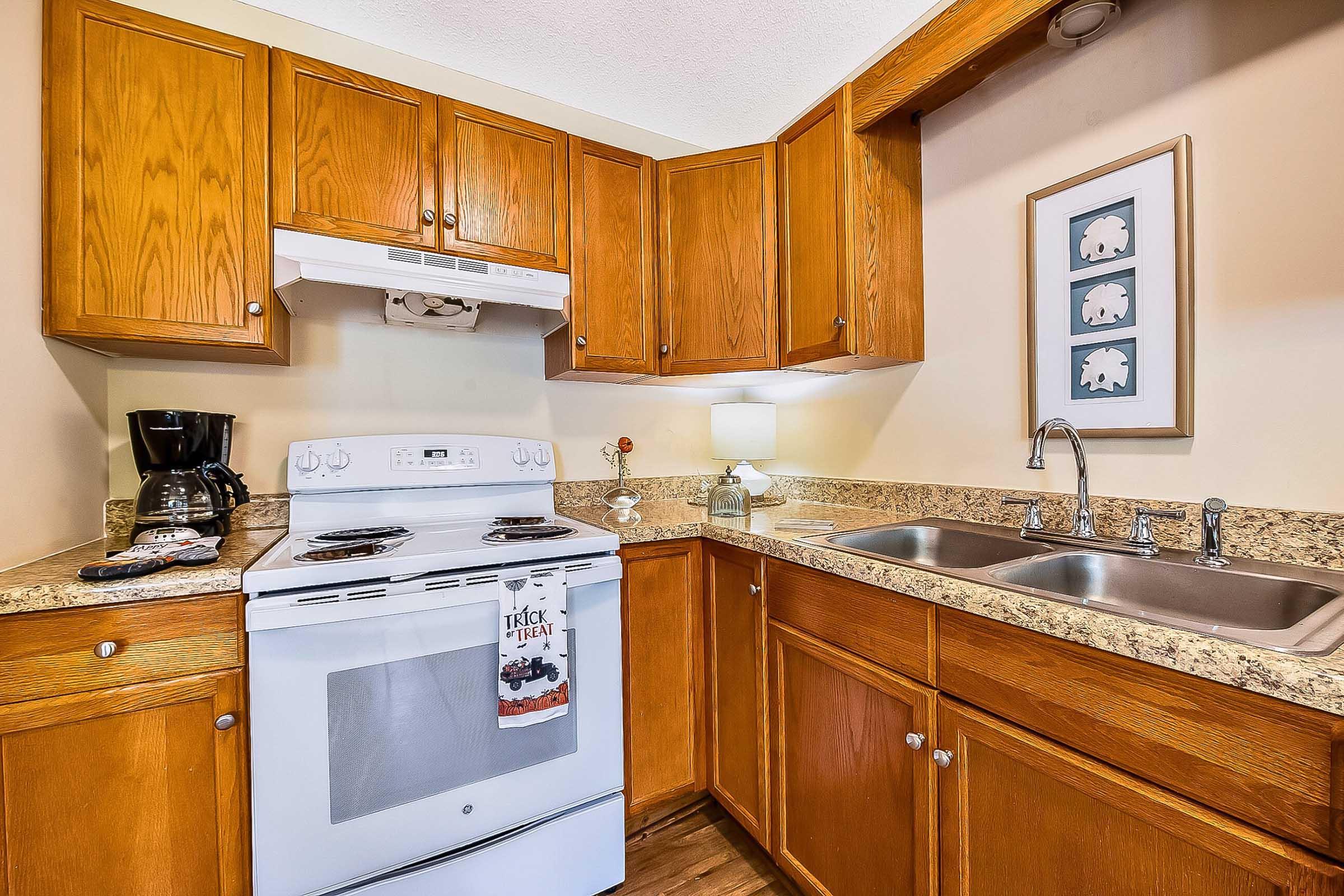 A cozy kitchen with wooden cabinets, a white stove, and a double sink. The countertop features a coffee maker and a decorative lamp. A framed artwork is hanging on the wall, and there are towels on the stove. Warm lighting creates an inviting atmosphere.