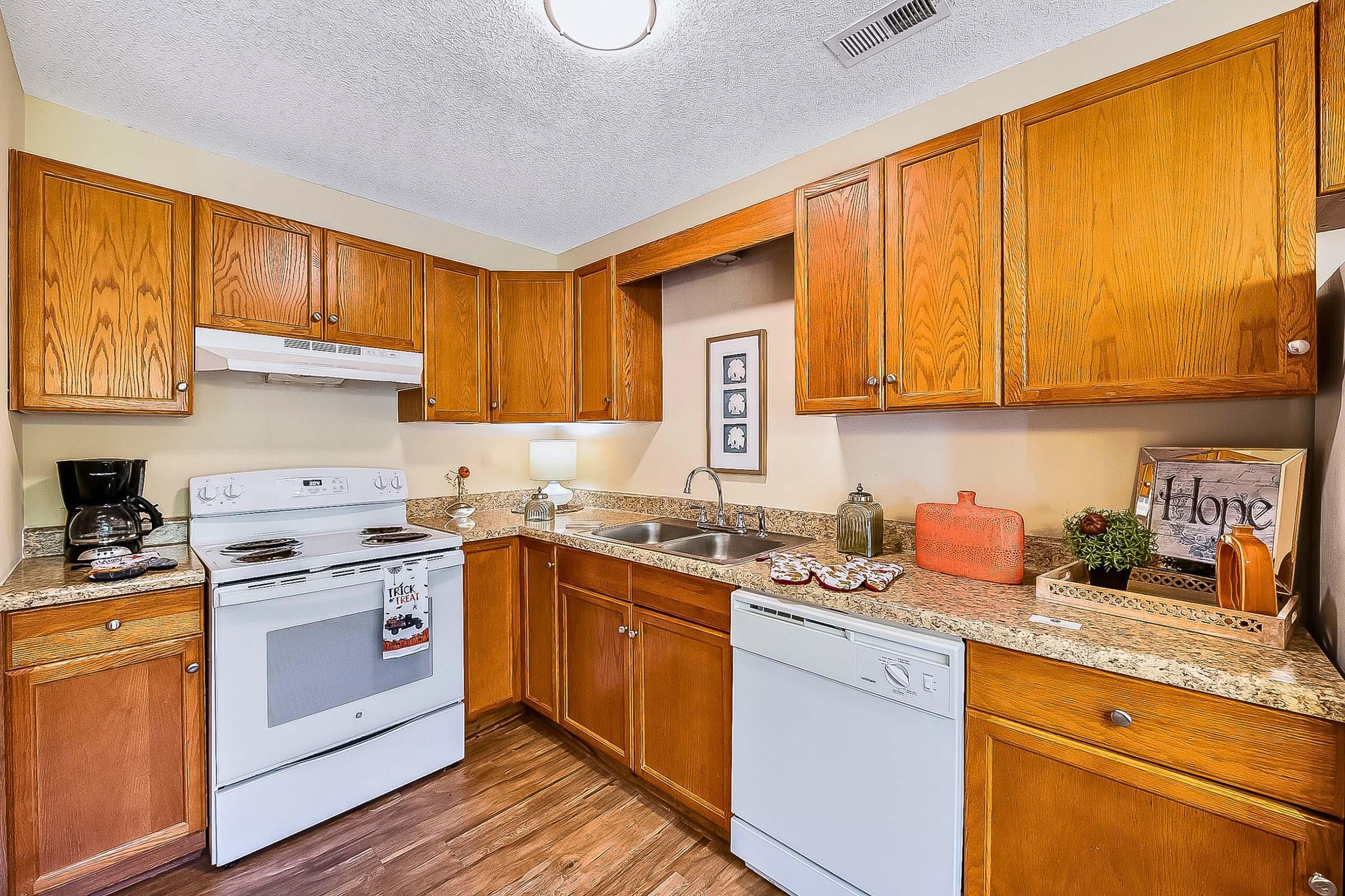 A well-designed kitchen featuring wooden cabinets, a white stove and dishwasher, a granite countertop, a sink with a lamp, and small decorative items like plants and a sign reading "Hope." The space has warm lighting and a cozy atmosphere.