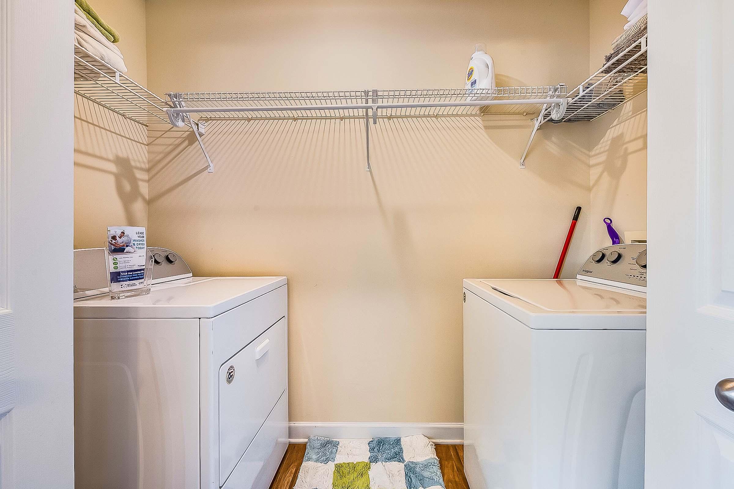 A spacious laundry room featuring a white washer and dryer, a shelf with cleaning supplies, and a colorful rug on the floor. The walls are a soft beige, creating a warm atmosphere. Light enters the room from a nearby door, enhancing the bright, clean look.