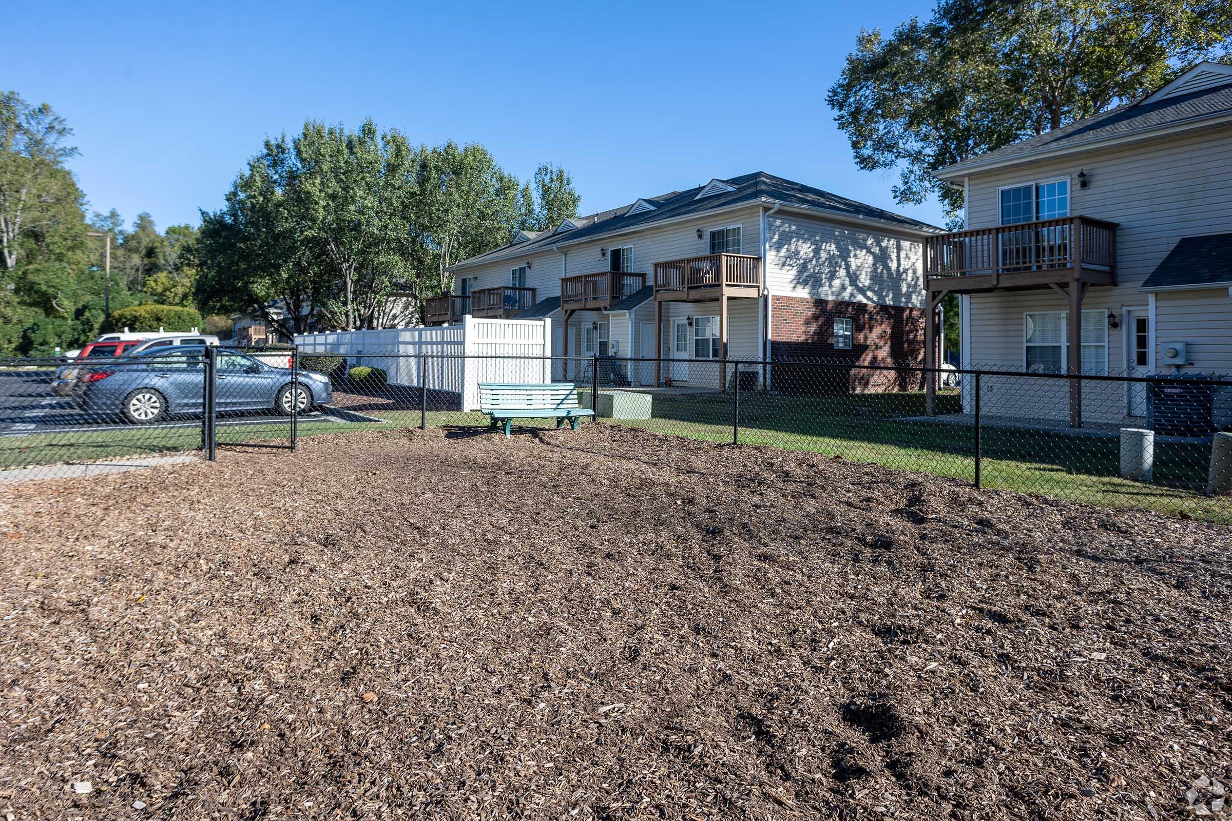 A fenced area with wood mulch, featuring a green bench. In the background, there are two multi-unit residential buildings and parked cars. The scene is set under a clear blue sky and surrounded by trees, creating a calm residential atmosphere.