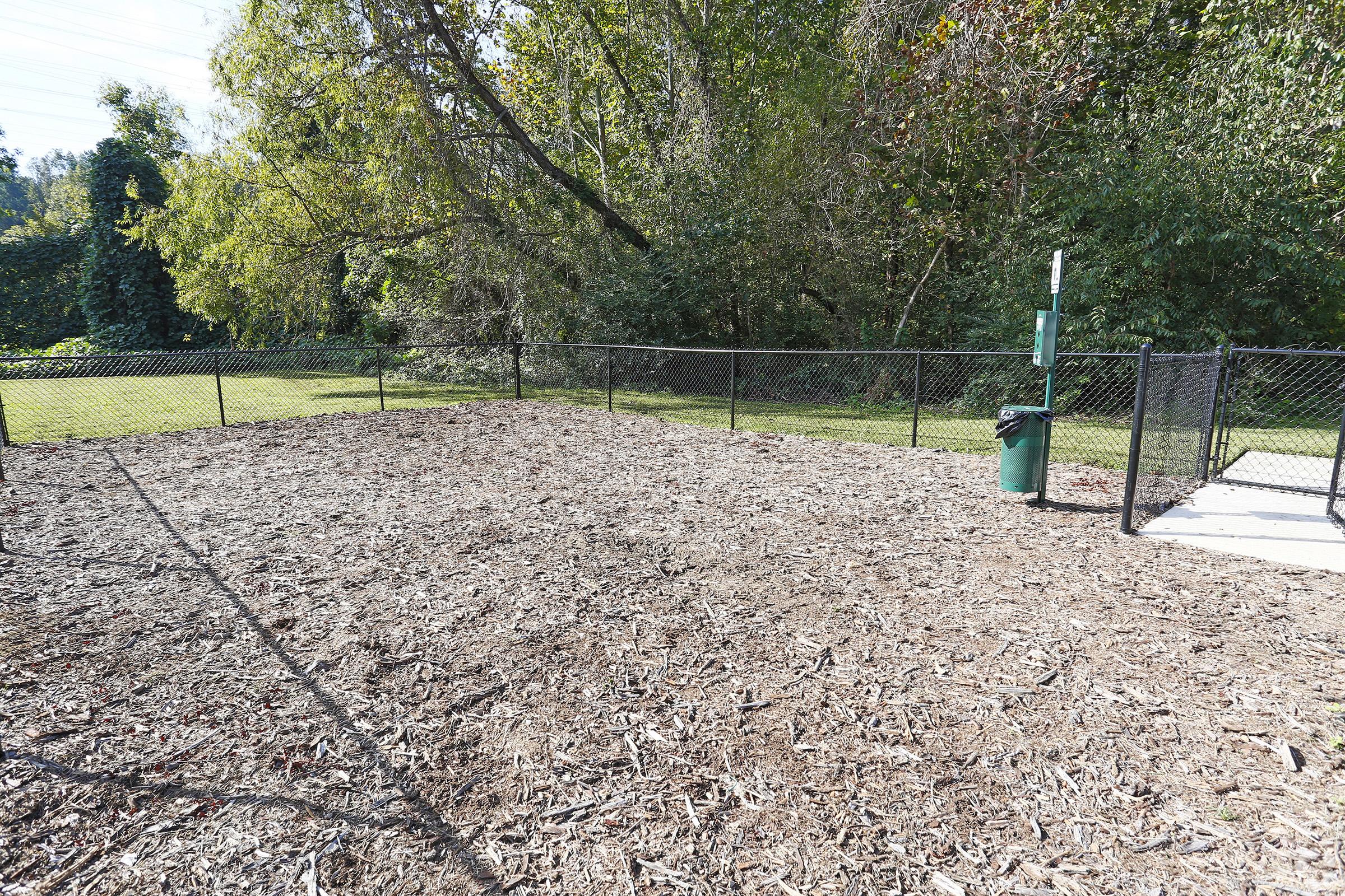 A fenced area with a woodchip-covered ground, surrounded by greenery. There is a green trash bin near a corner, and an open space ideal for activities or a dog park. The background features trees and shrubs, creating a natural setting.