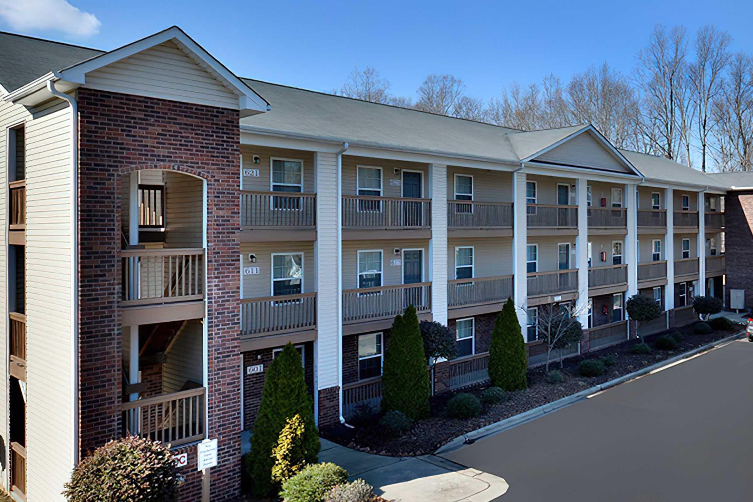 A multi-story apartment building with a brick and vinyl exterior. Balconies are visible on each floor, and there are neatly landscaped areas with shrubs and small trees. The sky is clear, indicating good weather, and the parking lot in front is well-maintained.