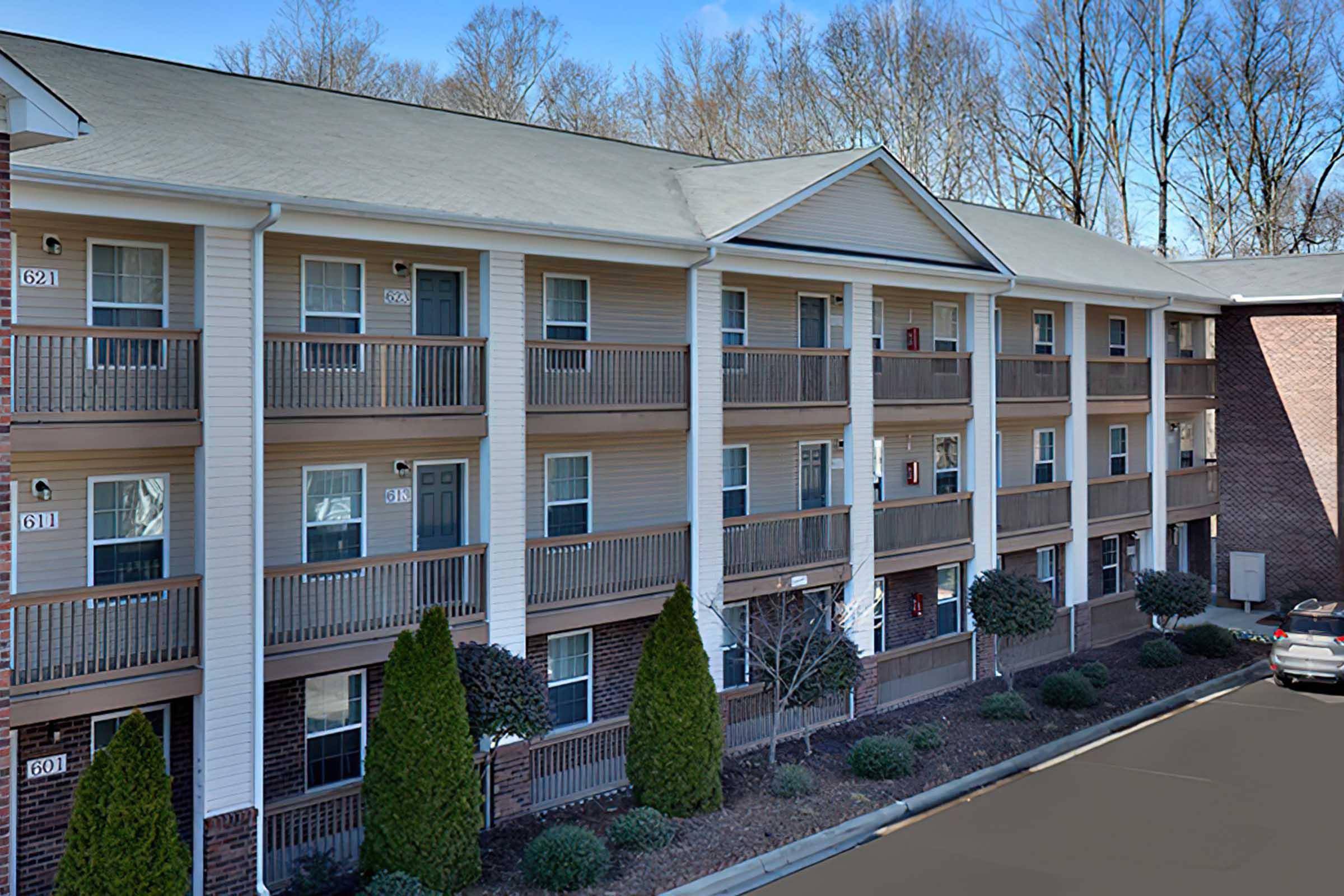 A multi-story apartment building featuring several balconies, with trees and shrubs lining the walkway. The building is designed in a traditional style with light-colored siding and brick accents. Parking is visible in the foreground, and the surrounding area is wooded.