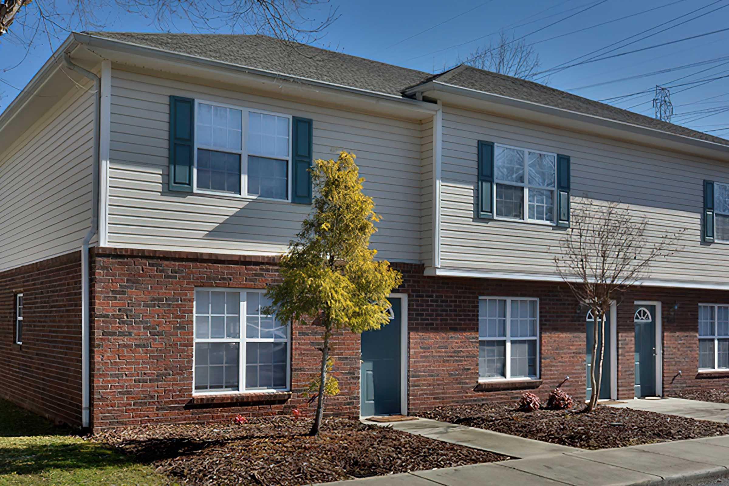 A two-story residential building featuring a mix of brick and siding exterior. The ground level has a small landscaped area with a young tree and flower beds. Windows are decorated with white frames and green shutters. Sidewalks lead up to the individual unit doors.