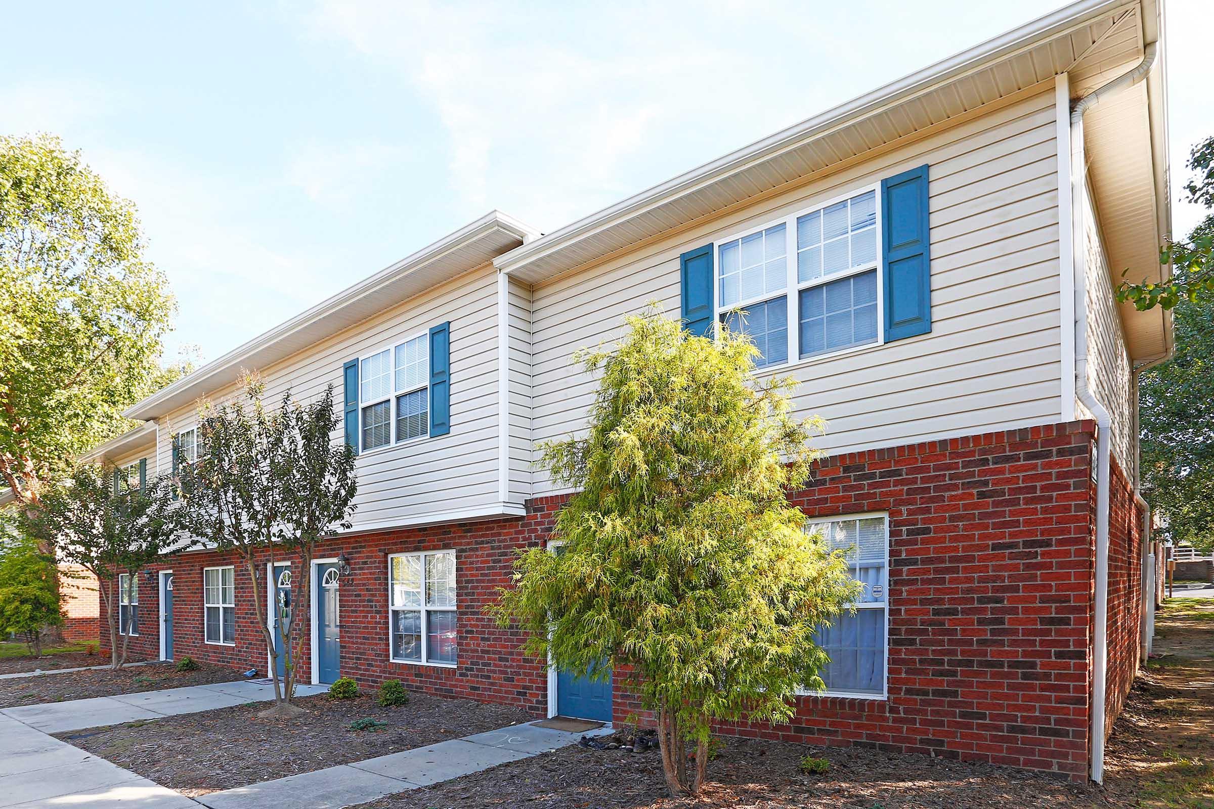 Two-story residential building with beige siding and red brick accents. Each unit has blue shutters and front doors. The landscaped area features small trees and shrubs, with a concrete walkway leading to the entrance. Natural light highlights the building's exterior on a clear day.